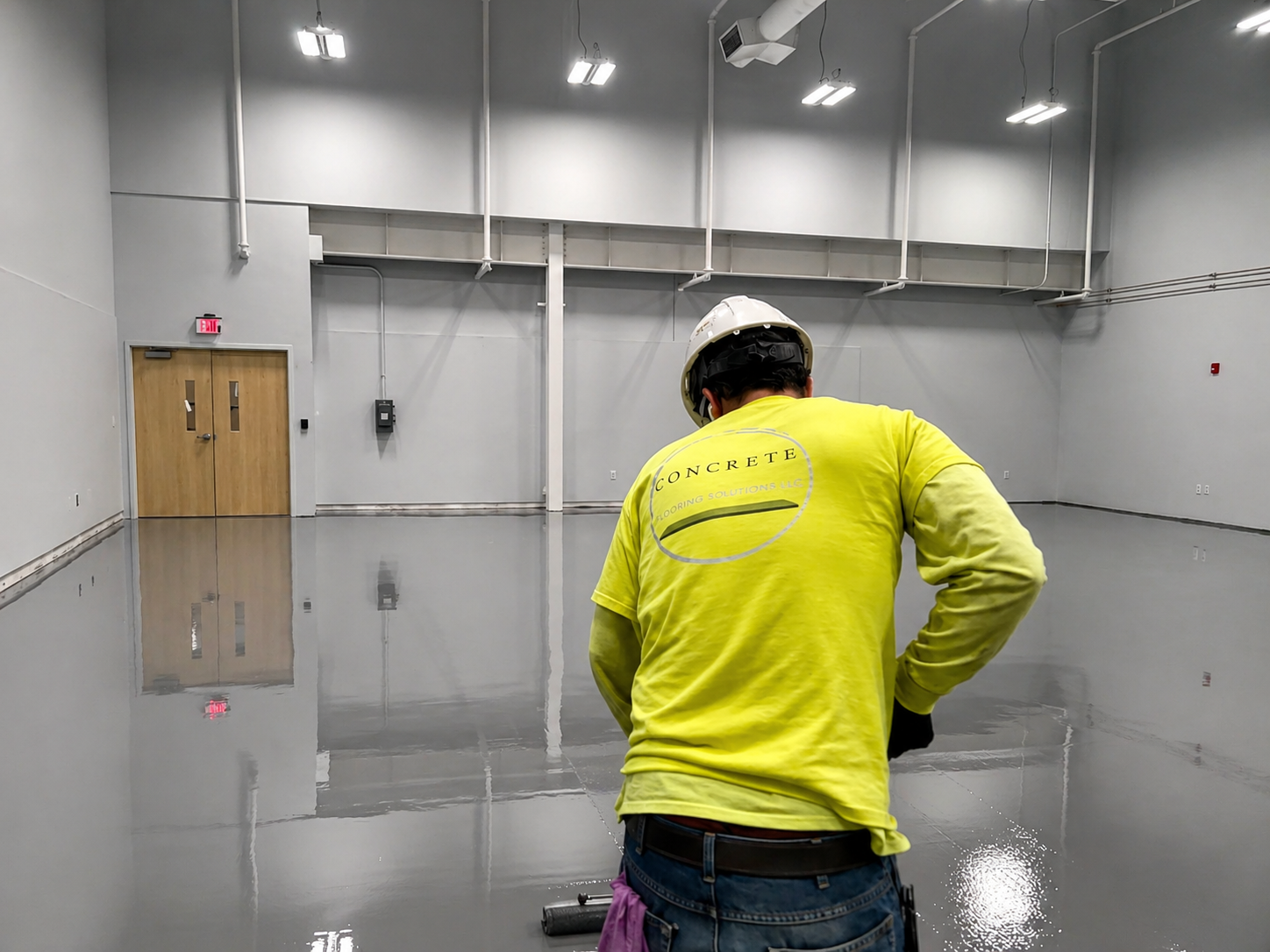 A worker wearing a yellow shirt with 'CONCRETE FLOORING SOLUTIONS LLC' on the back, a helmet, and gloves applying a gray epoxy coating on a large indoor concrete floor. The room has white walls, ceiling lights, and a closed wooden door.