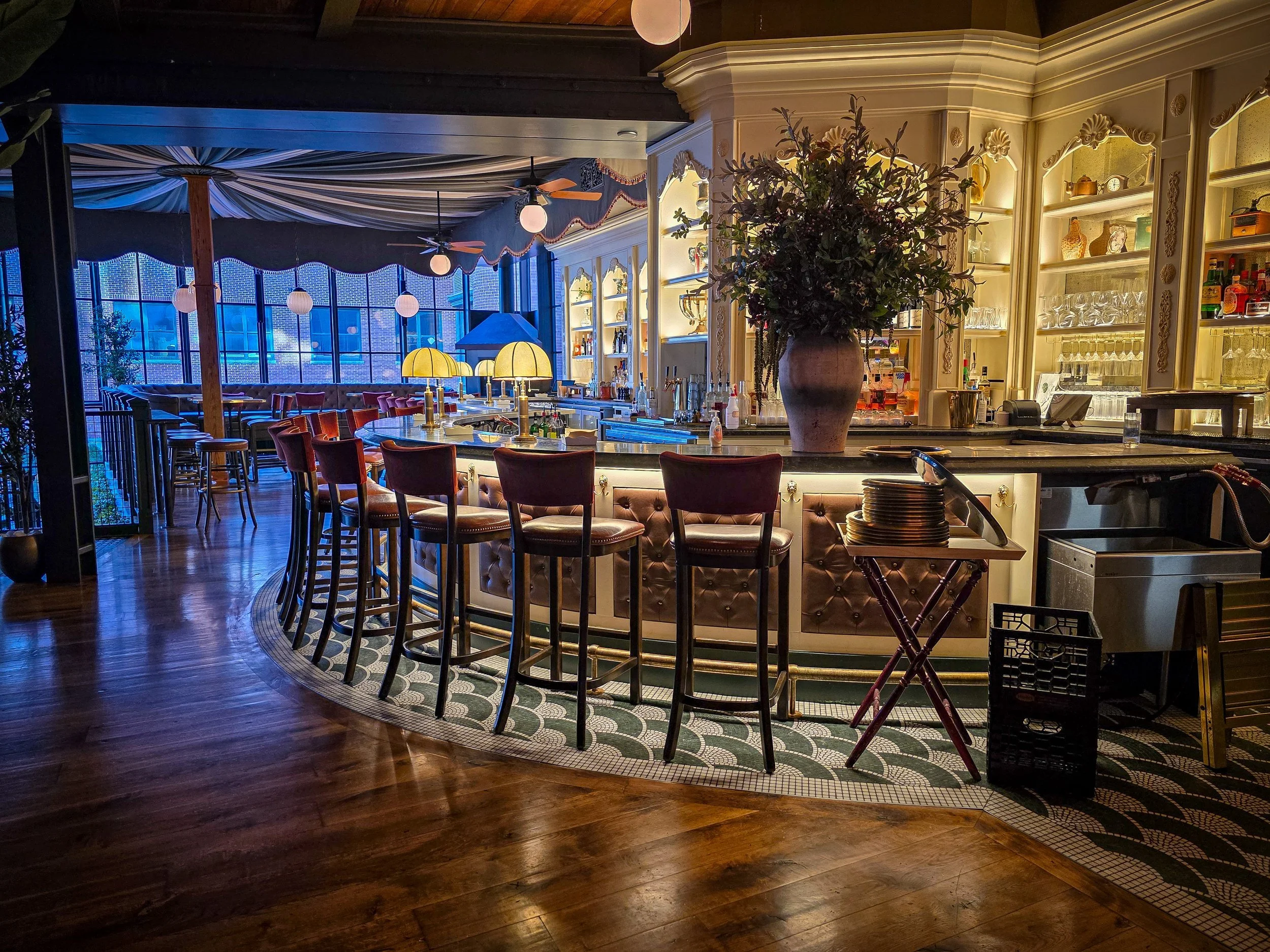 A bar interior with a semi-circular counter, bar stools, large floral arrangement, illuminated shelves with glassware and bottles, and a ceiling with draped fabric and hanging lights, nighttime visible through large windows.