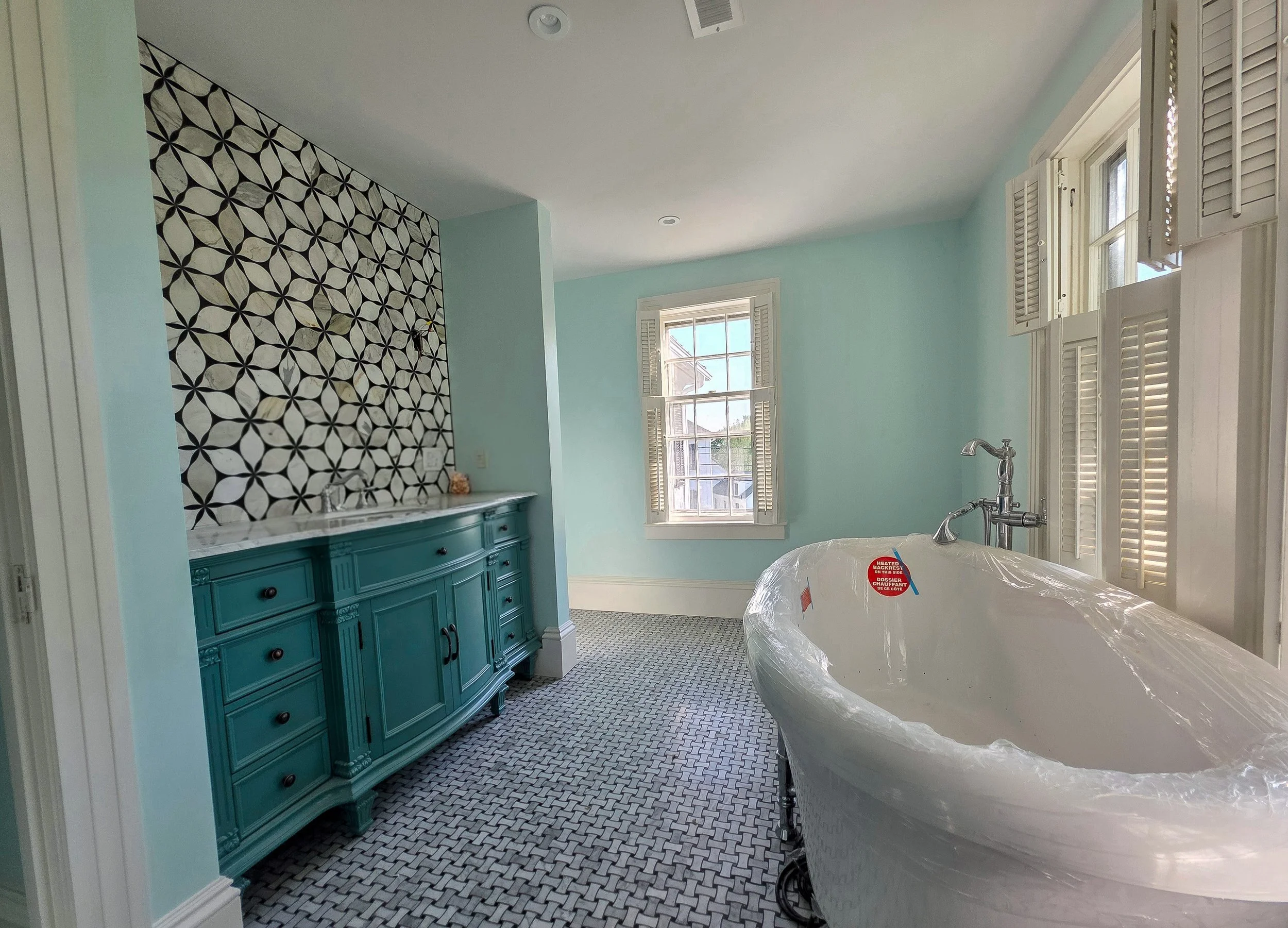 A bathroom under renovation with a blue vanity, patterned tile flooring, and a bathtub covered with plastic. There's a large window with white shutters, and one wall features black and white geometric patterned wallpaper.