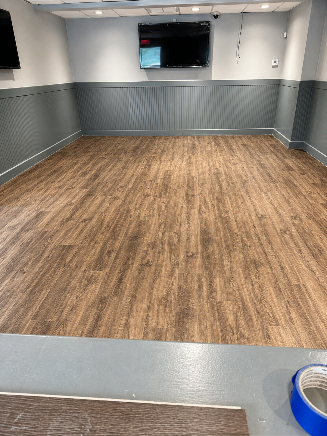 Empty room with wood flooring, gray walls, two mounted TVs, and a ceiling with recessed lights.