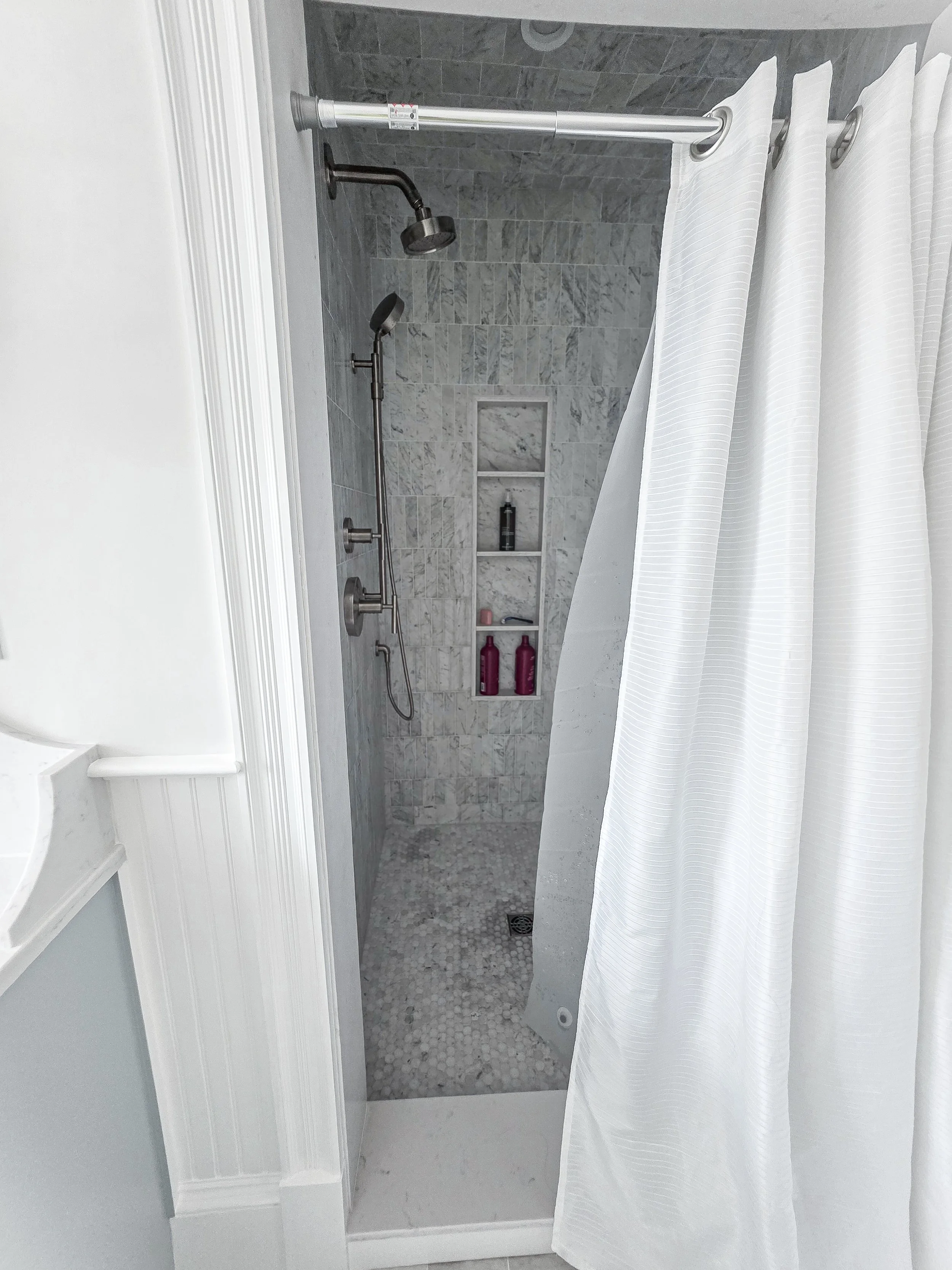 A bathroom shower with gray marble tiles, a built-in niche holding toiletries, a white shower curtain, and various shower fixtures including a handheld showerhead.