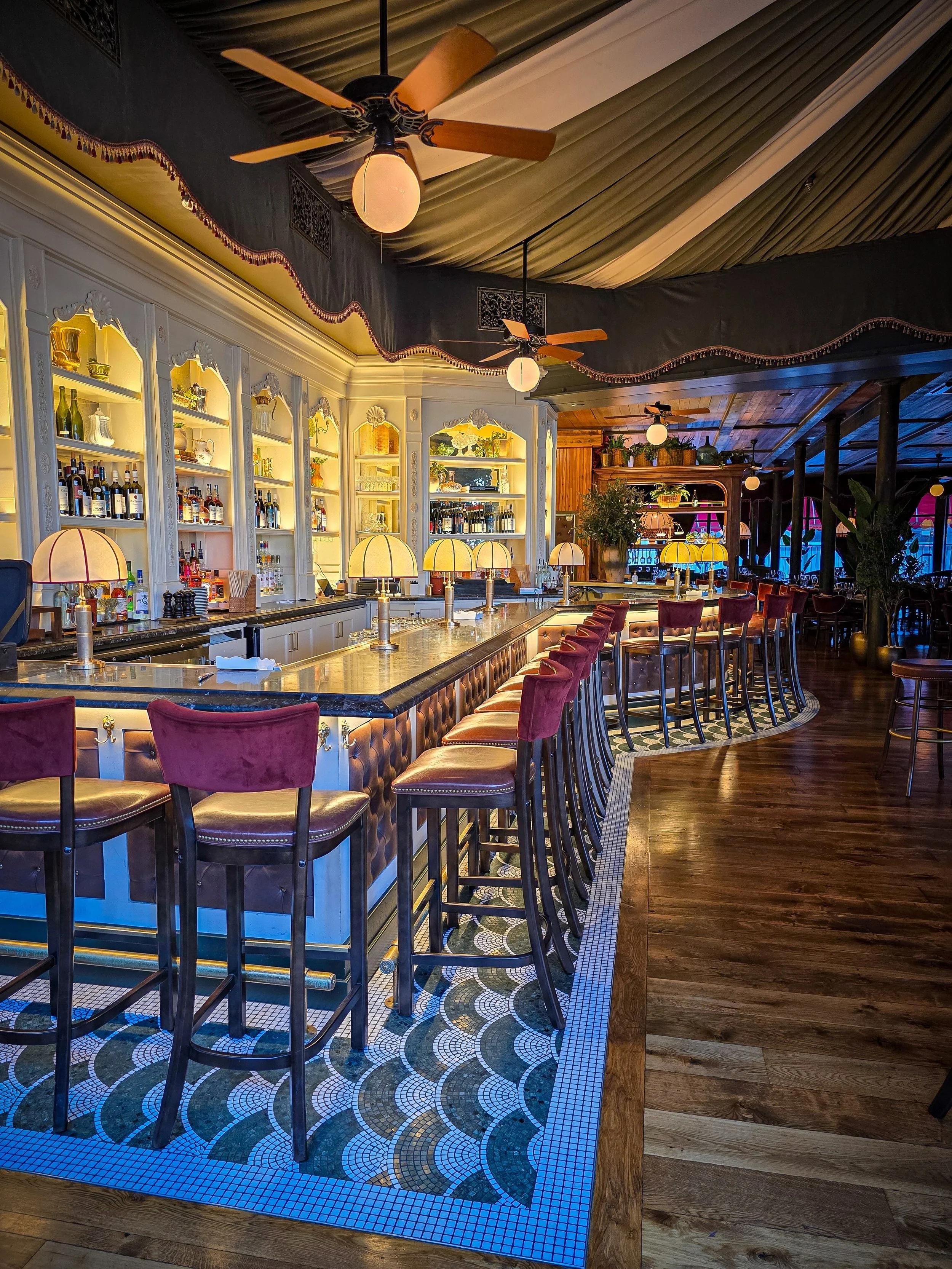 Interior of a bar with a marble counter, red velvet barstools, decorative lamps, a white ornate shelf with bottles, and ceiling fans with wooden blades, wood and tile flooring, and a dark draped ceiling.