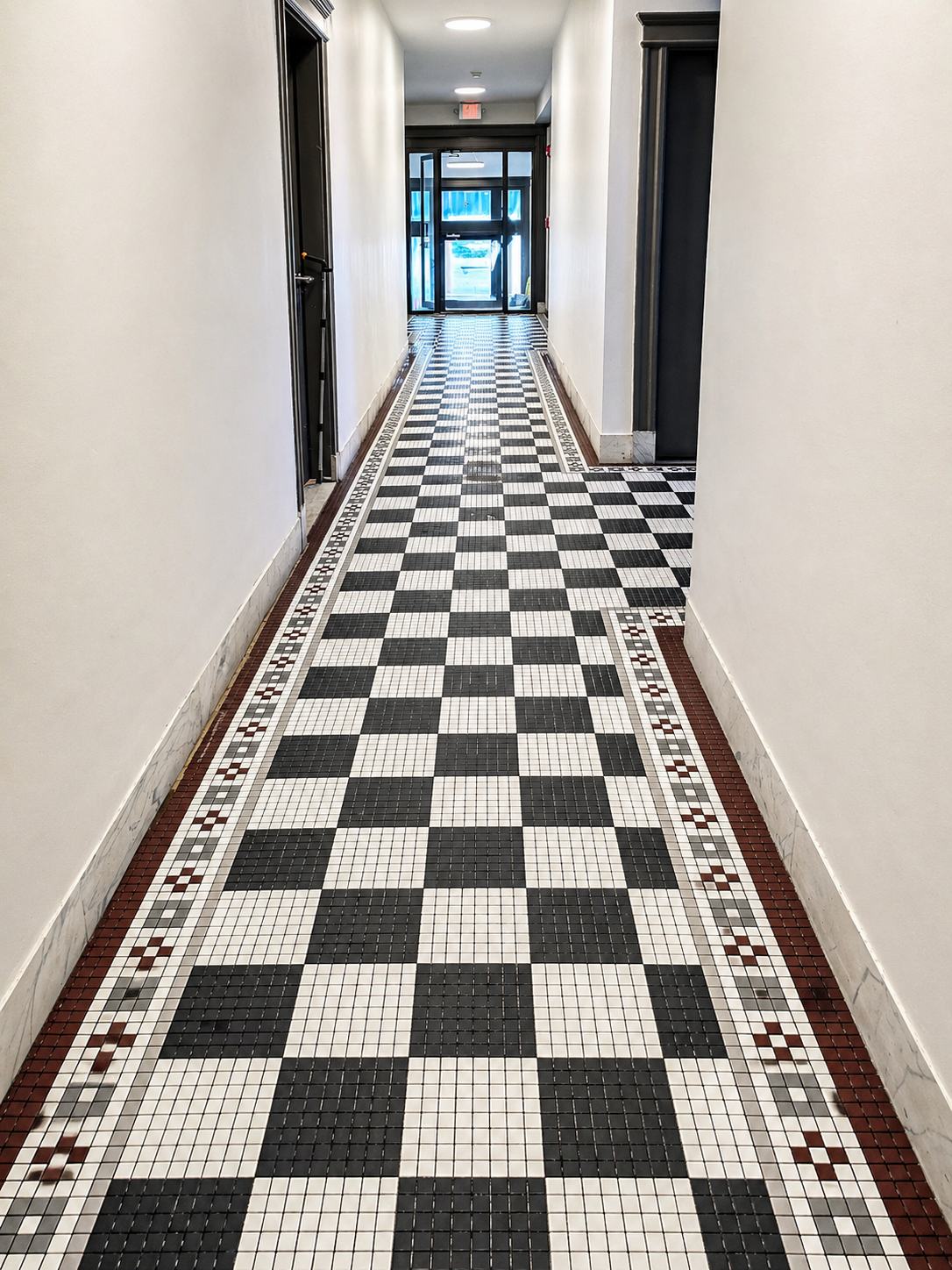Long corridor with black, white, and red checkered mosaic tile floor, white walls, and dark door frames, leading to an entrance with glass doors.