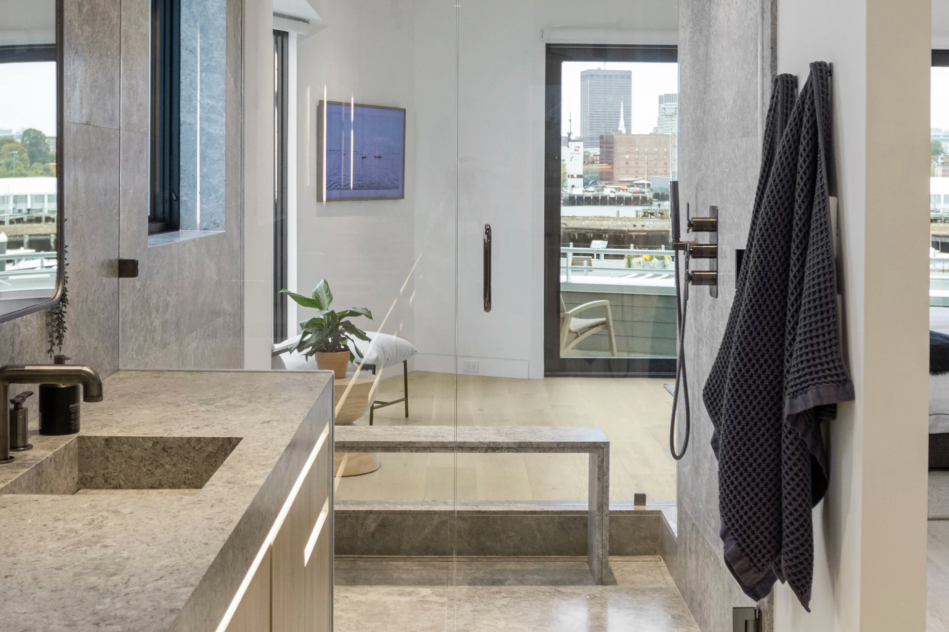 Modern bathroom with a view of a city skyline from a balcony. Features a marble countertop, a small potted plant, a large mirror, and black towels hanging on the wall.