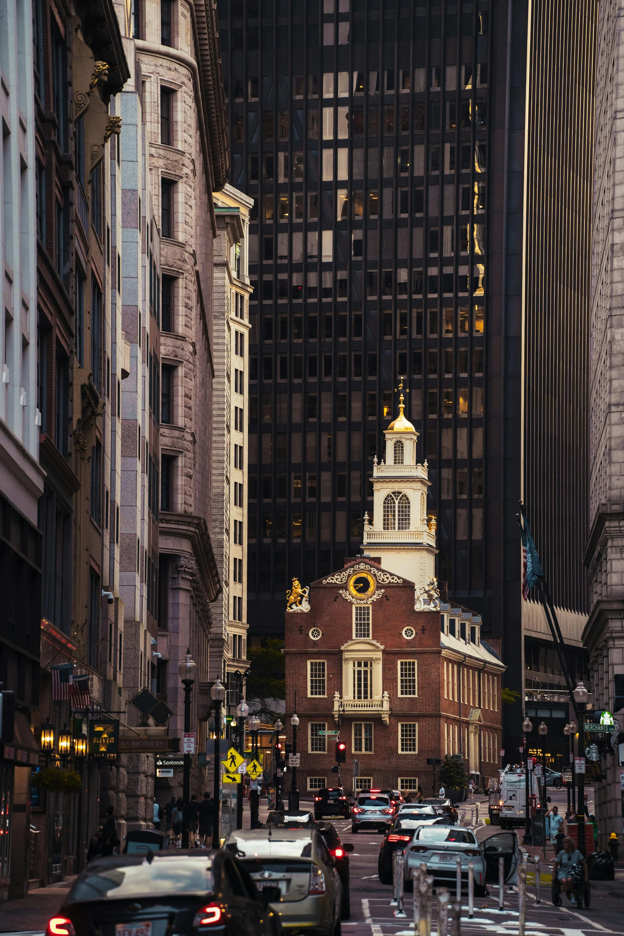 Street view in a city with historic and modern buildings, cars on the road, pedestrians, and a historic building with a clock and gold decoration at the center of the image.