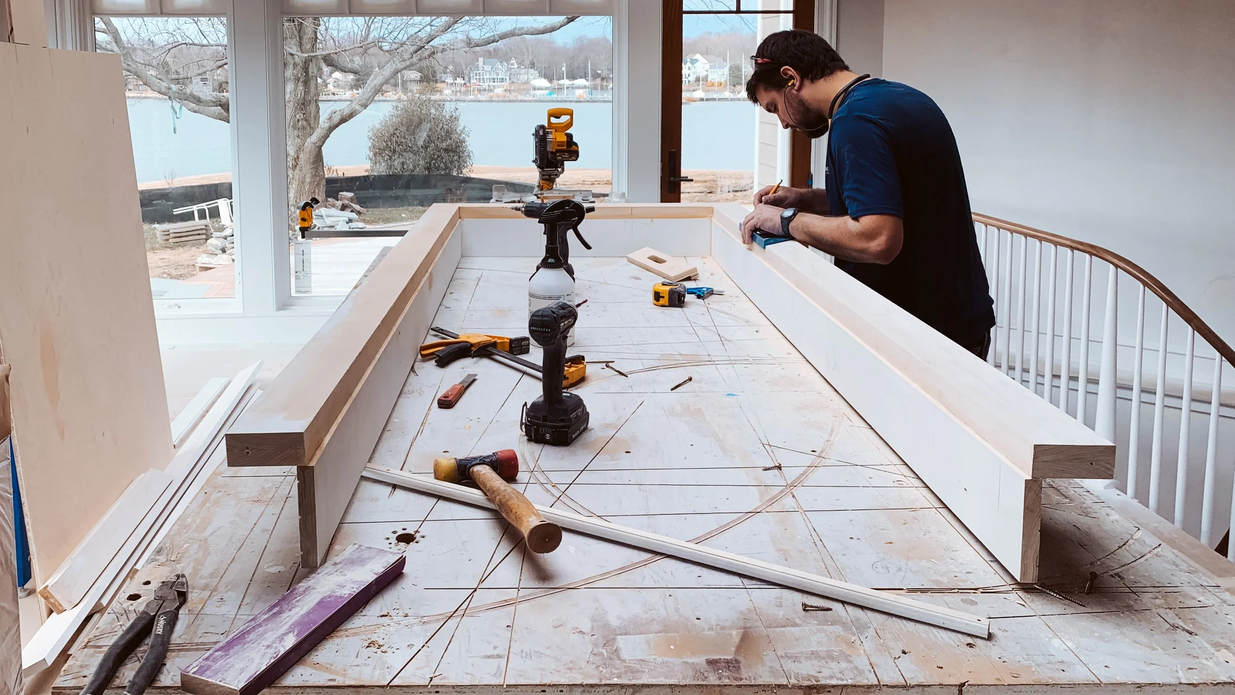 A man working on a woodworking project at a large window with a view of a lake and trees. The workspace has tools, wood pieces, and construction materials on a table.
