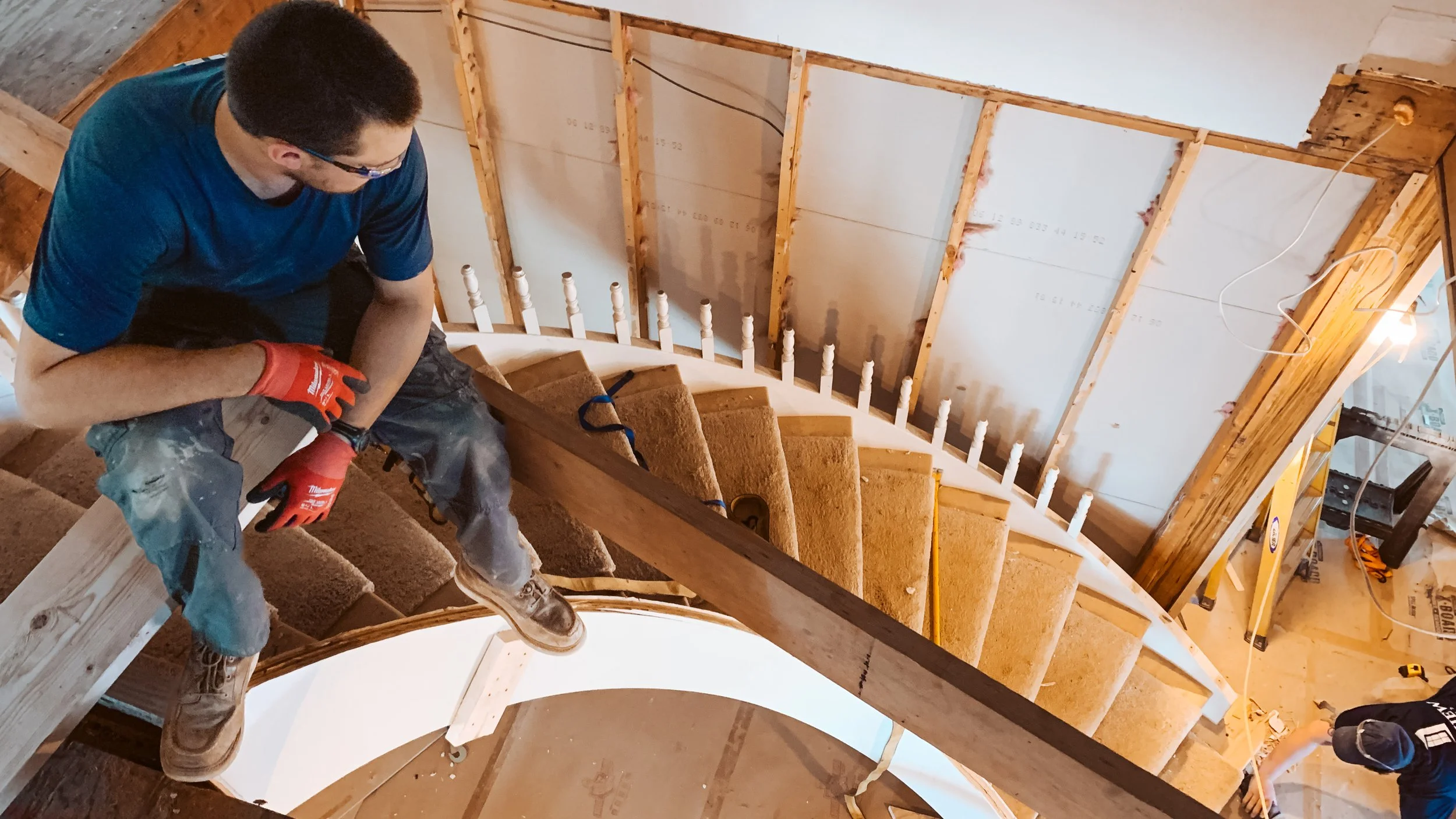 A man in a blue shirt and red gloves working on a staircase under construction, with exposed wooden framing and insulation visible on the walls.