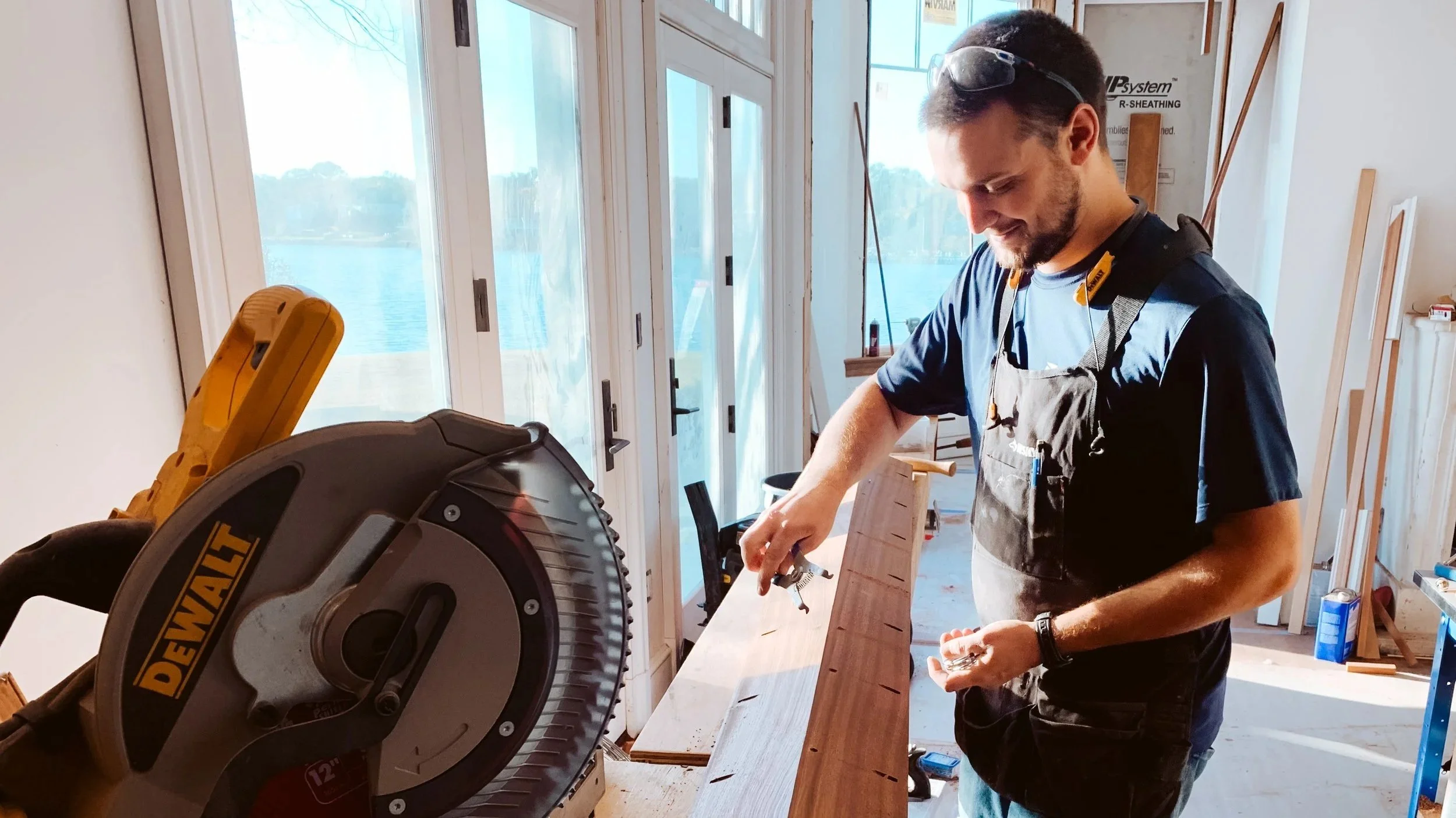 A man using a circular saw to cut a wooden board inside a room near large windows, with construction materials and tools in the background.