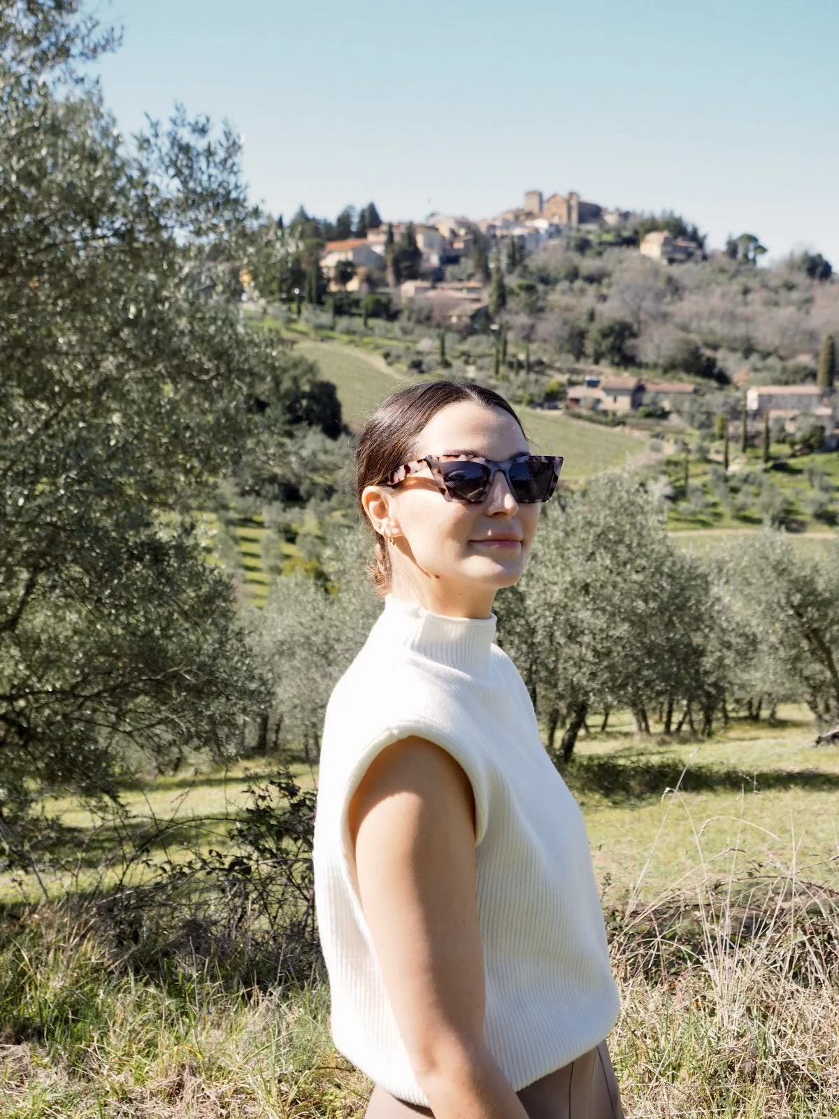 A woman with dark brown hair wearing sunglasses and a cream sleeveless sweater stands outdoors in a green Italian hillside landscape with trees and a distant hillside town under a clear blue sky.