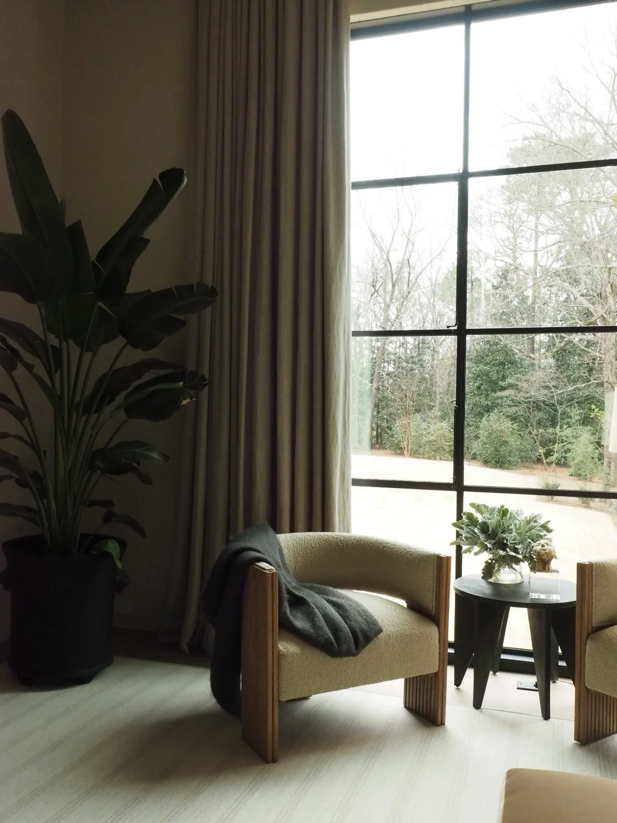 A corner of a room with beige chairs, a small dark table with a floral arrangement, large window with curtains, and a tall plant with large dark leaves.