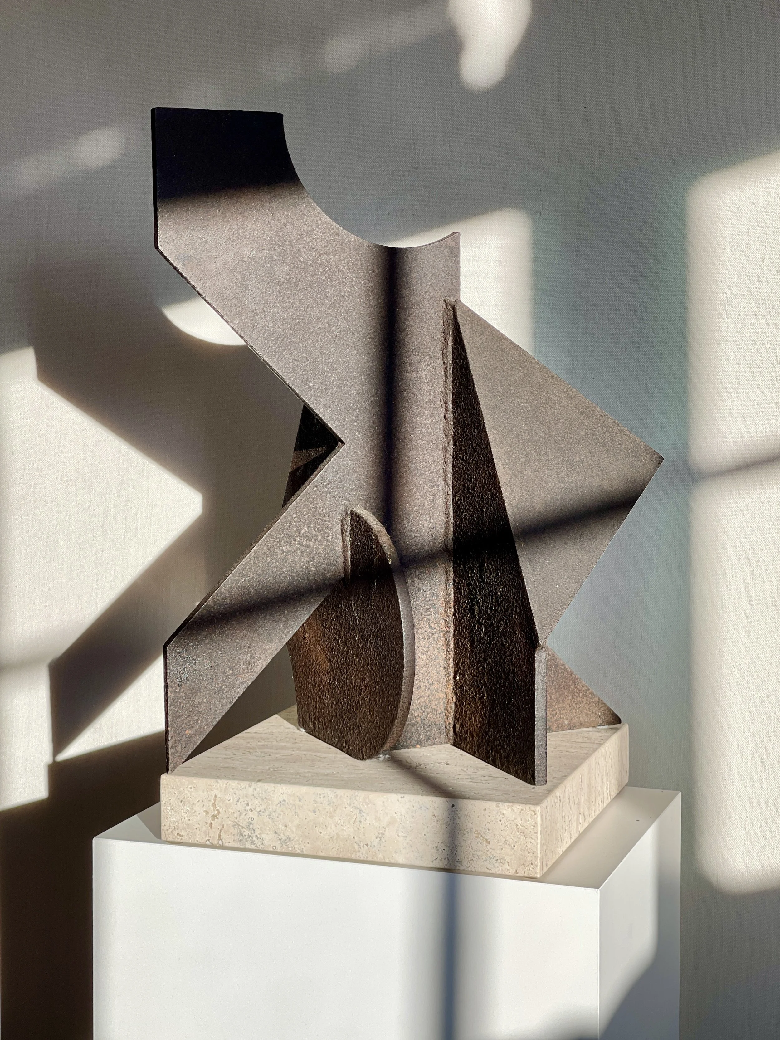 Abstract geometric sculpture made of weathered metal, mounted on a beige stone base and placed on a white pedestal, with shadows cast by natural light.