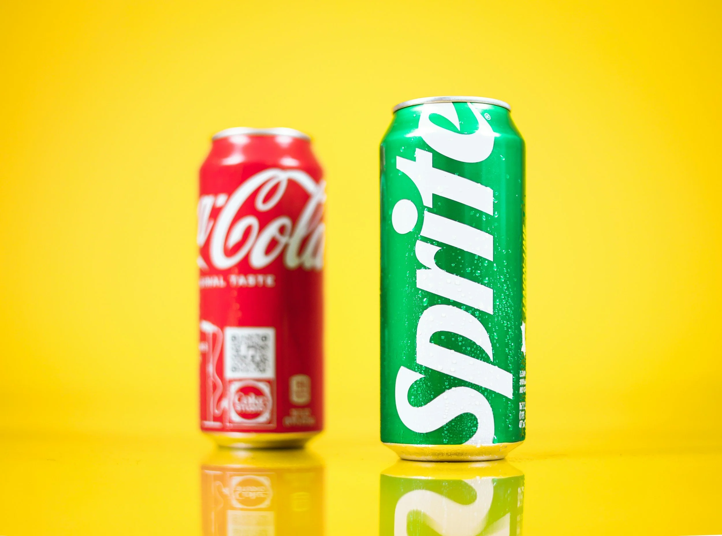 Close-up of a Sprite soda can with water droplets, positioned in front of a Coca-Cola can, both against a vibrant yellow background.