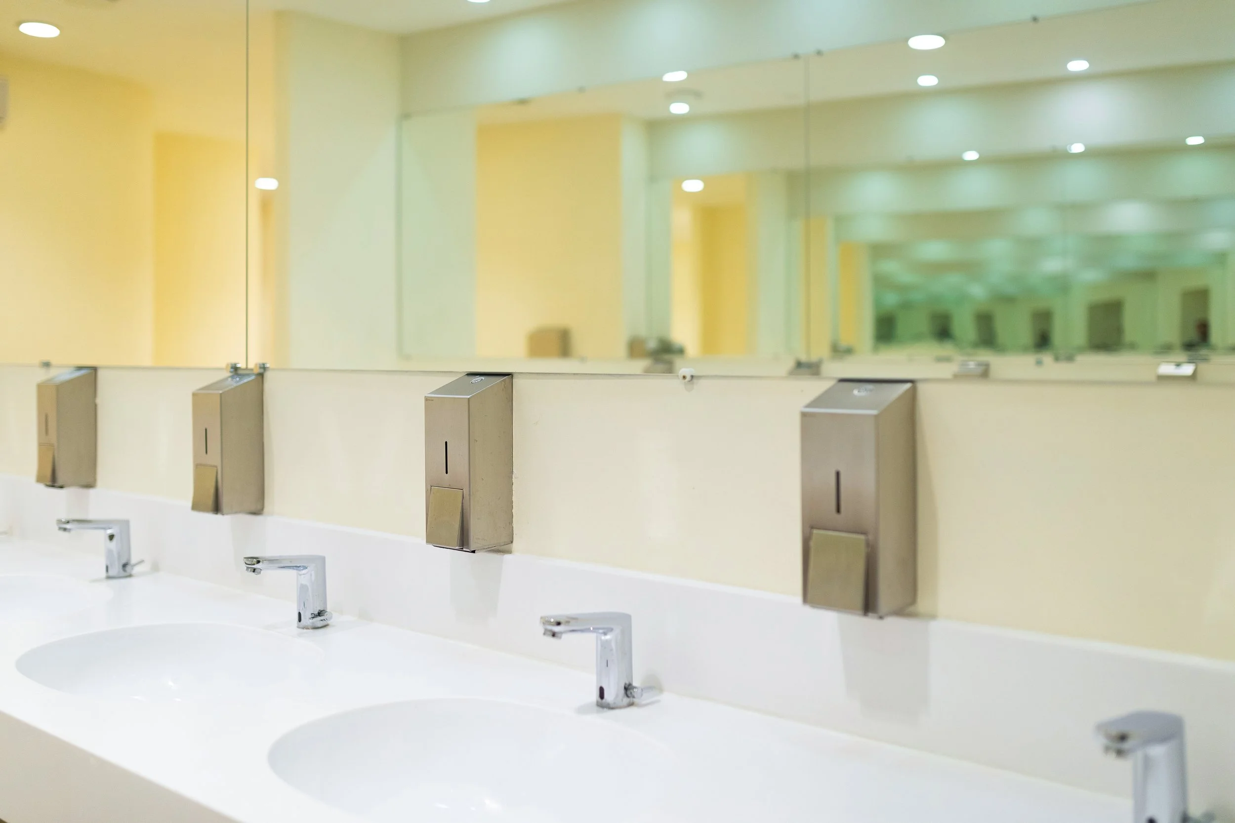 Public restroom with three sinks, silver soap dispensers, large mirror, and yellow walls with ceiling lights.