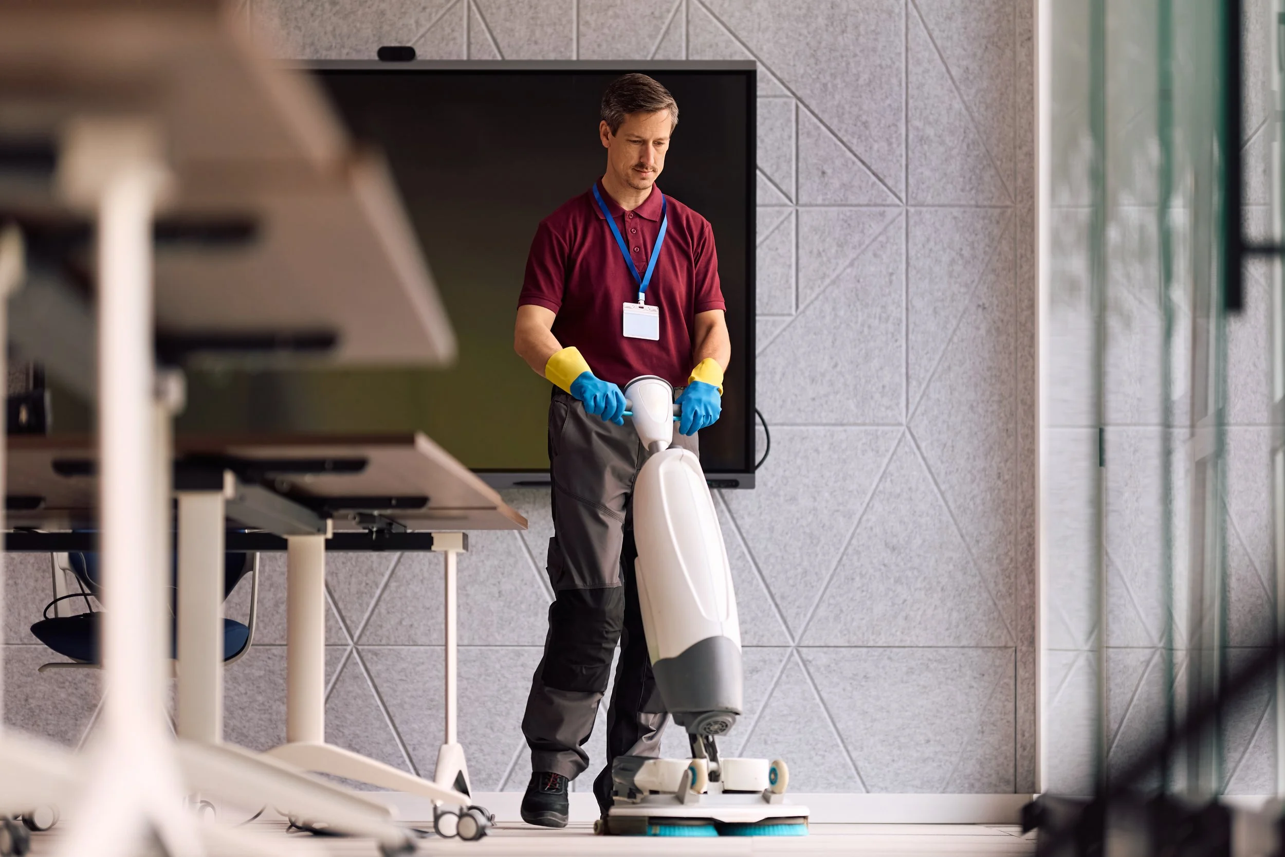 A man wearing a burgundy polo shirt, gray pants, yellow and blue gloves, and an ID badge on a blue lanyard is disinfecting a scooter with a handheld disinfectant device in an office or commercial space with tiled walls and glass windows.