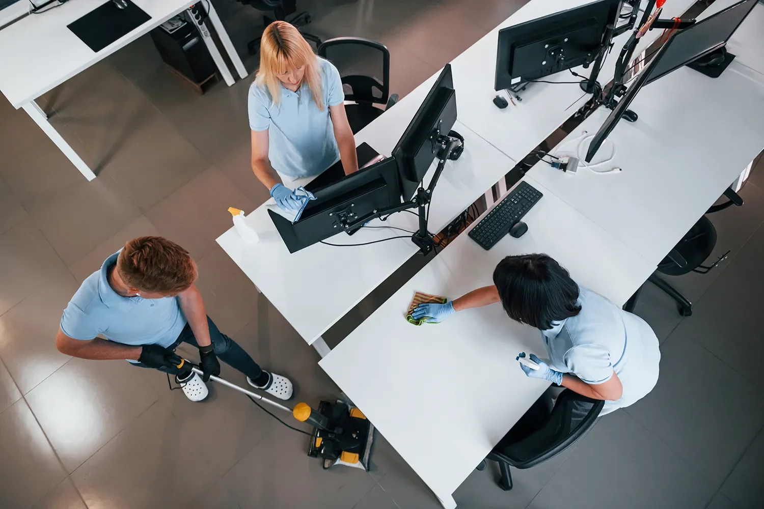 Three people cleaning an office. One person is mopping the floor, and two others are wiping down desks and monitors.