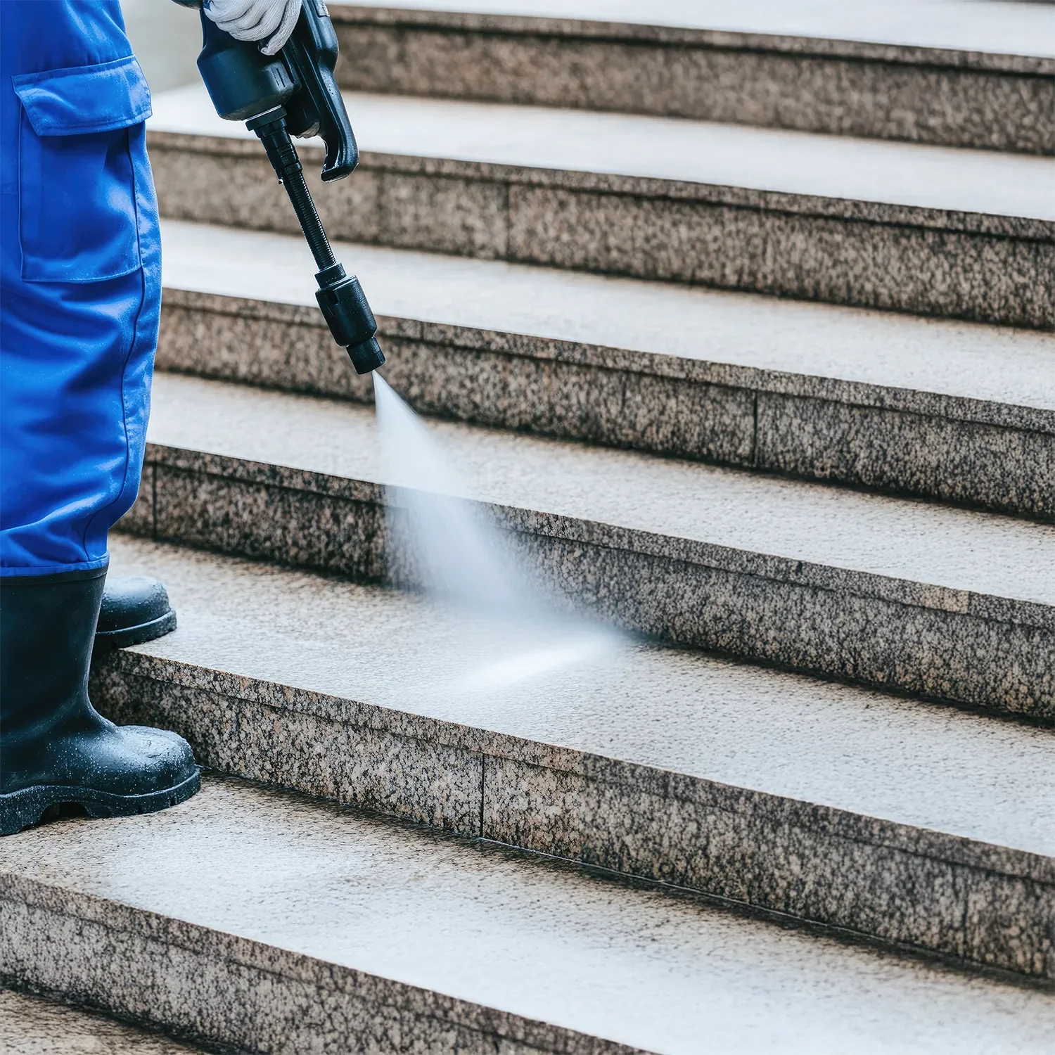A person spraying water on granite steps with a pressure washer. The person is wearing blue coveralls, black boots, and gray gloves.