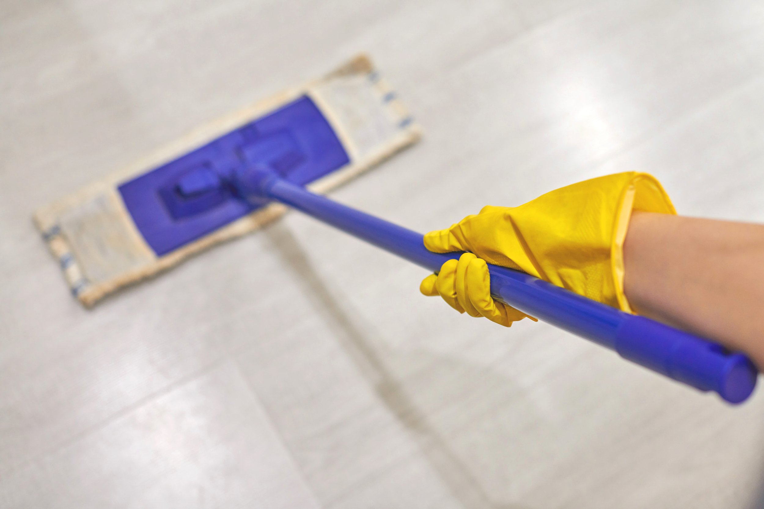 Person wearing yellow gloves mops the floor with a blue mop and a yellow mop bucket.