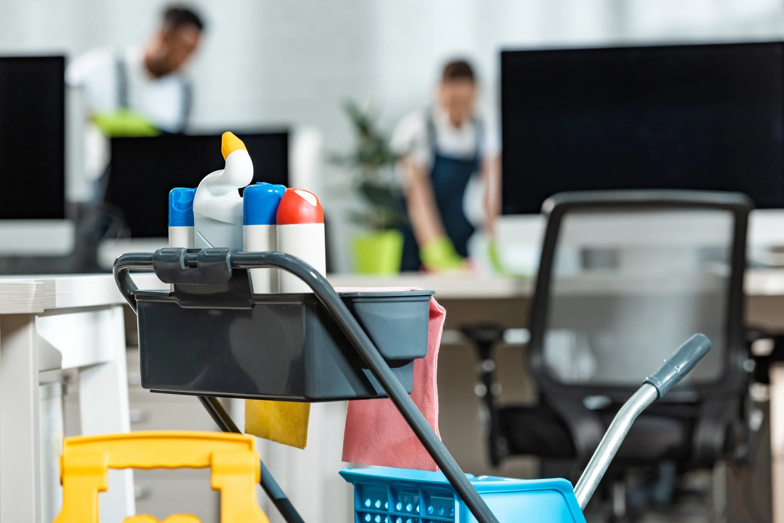 Cleaning supplies on a cart with two blurred janitors working in the background