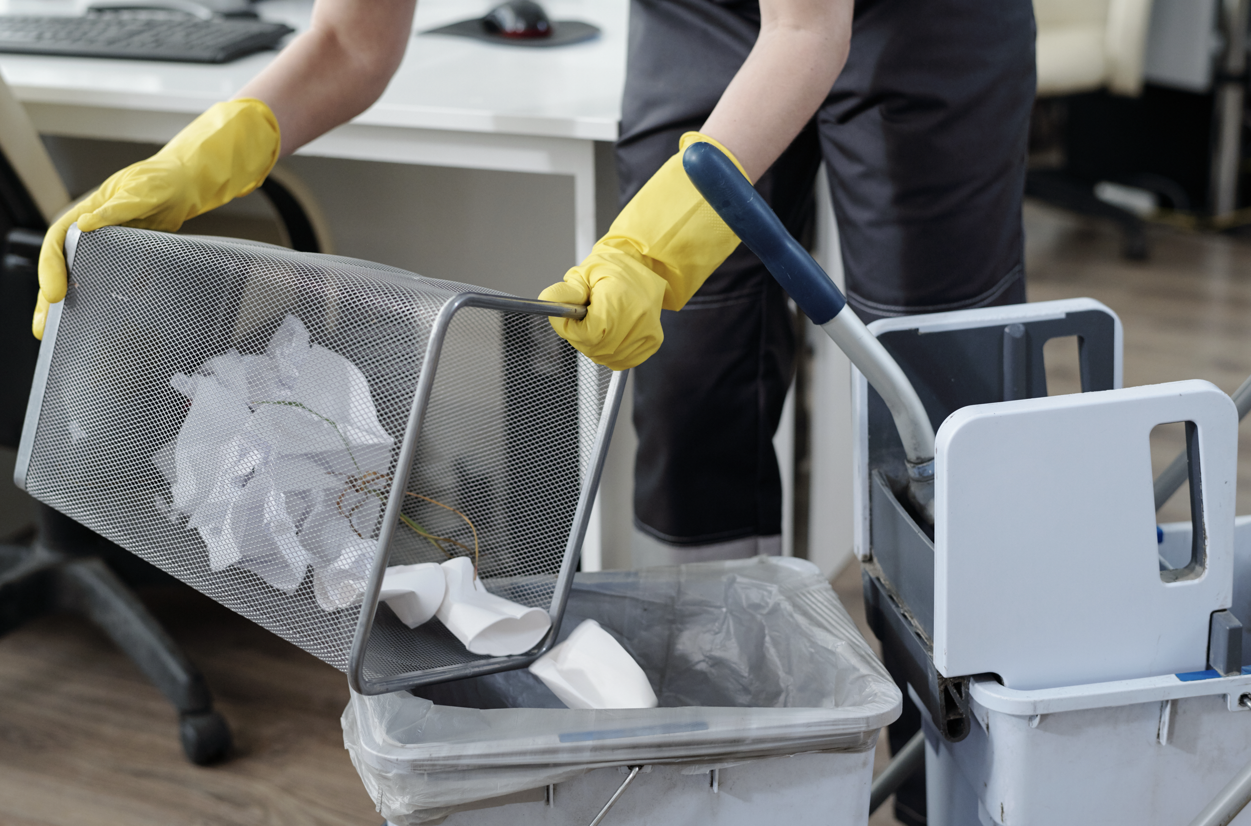 Person wearing yellow rubber gloves handling a mesh trash bin filled with paper and plastic items in an office or medical setting.