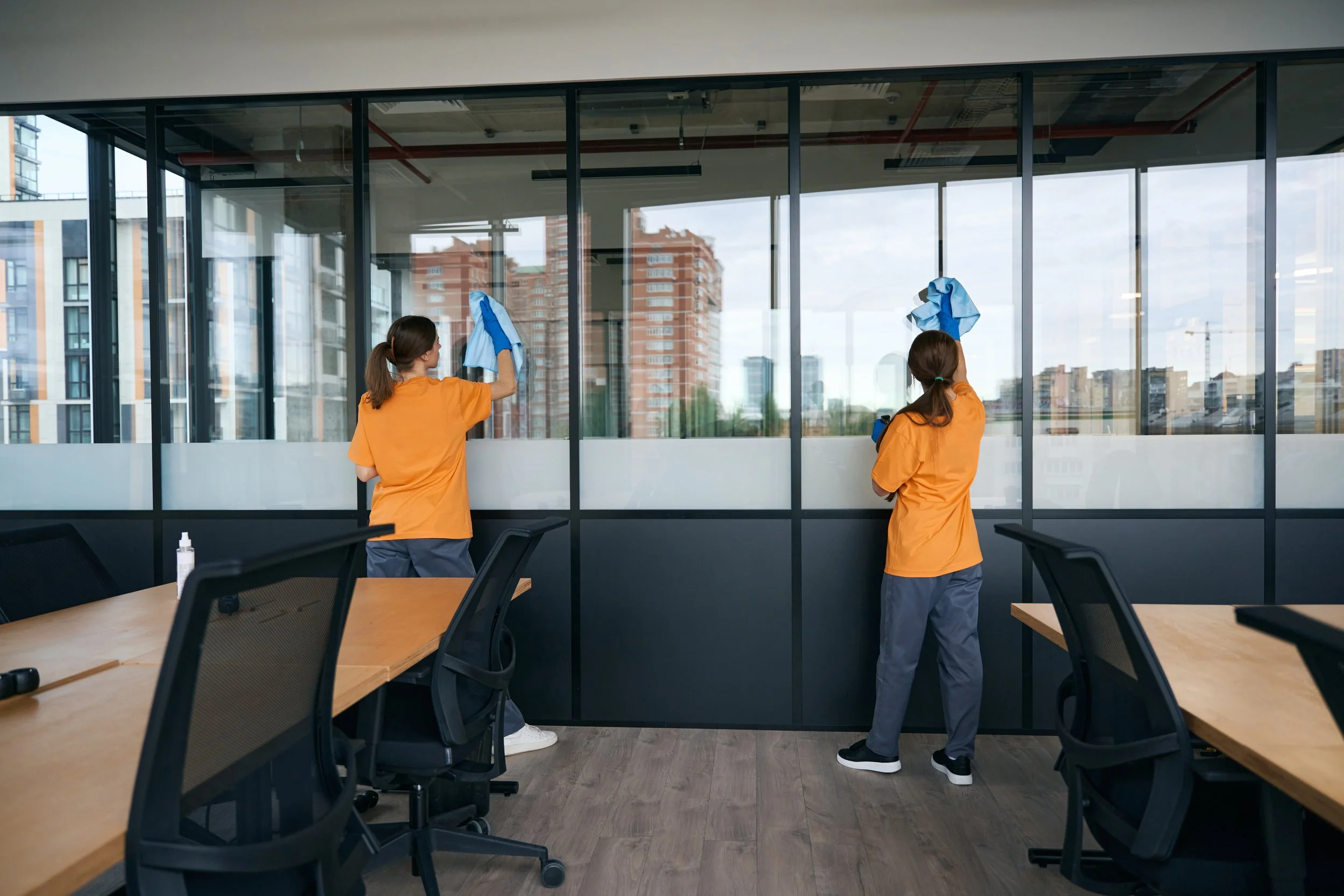 Two women in orange shirts and gray pants cleaning large glass windows in a modern office building, with city buildings visible outside.