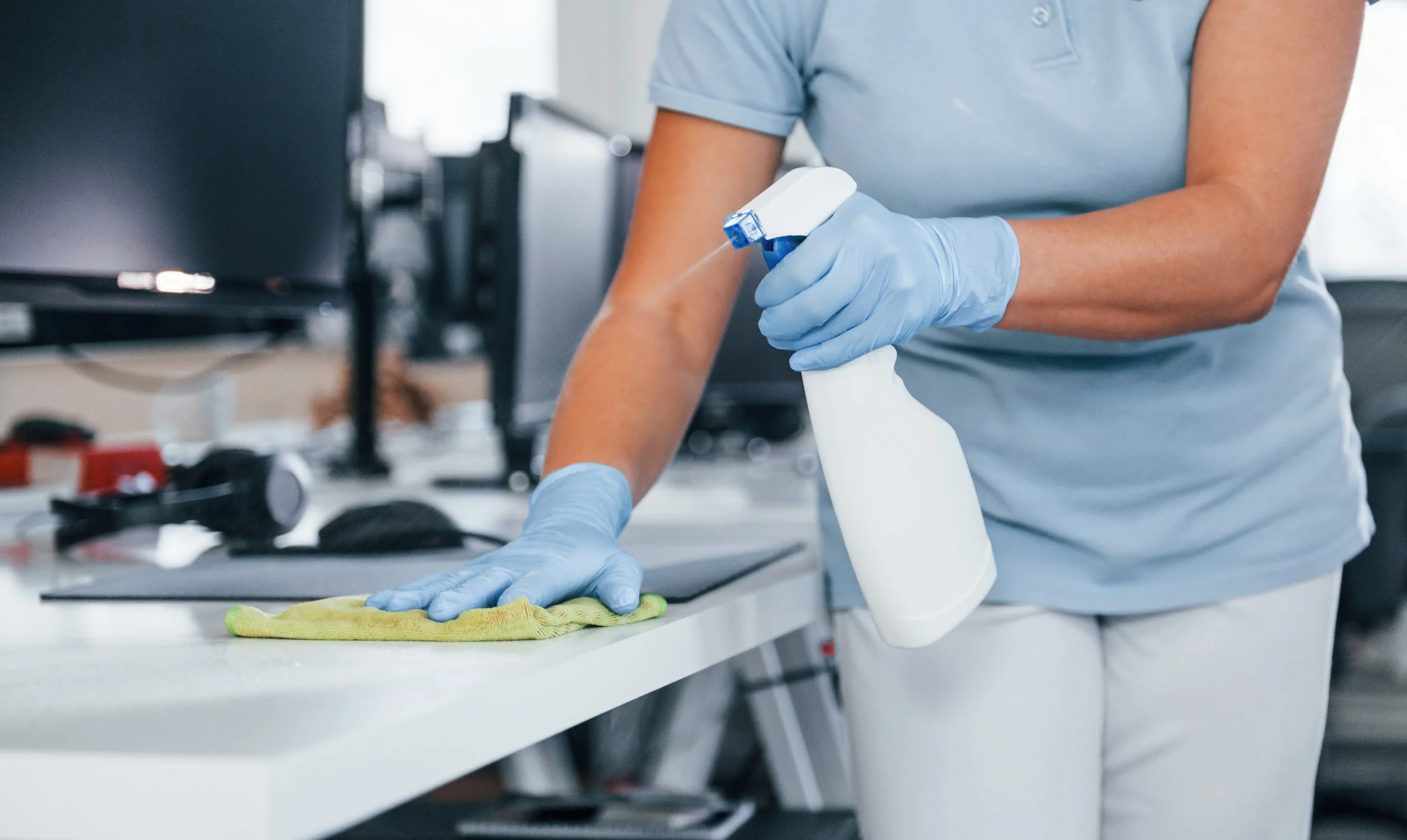 A person in a light blue shirt and blue gloves is cleaning a surface with a yellow cloth and a spray bottle in a laboratory or medical setting.