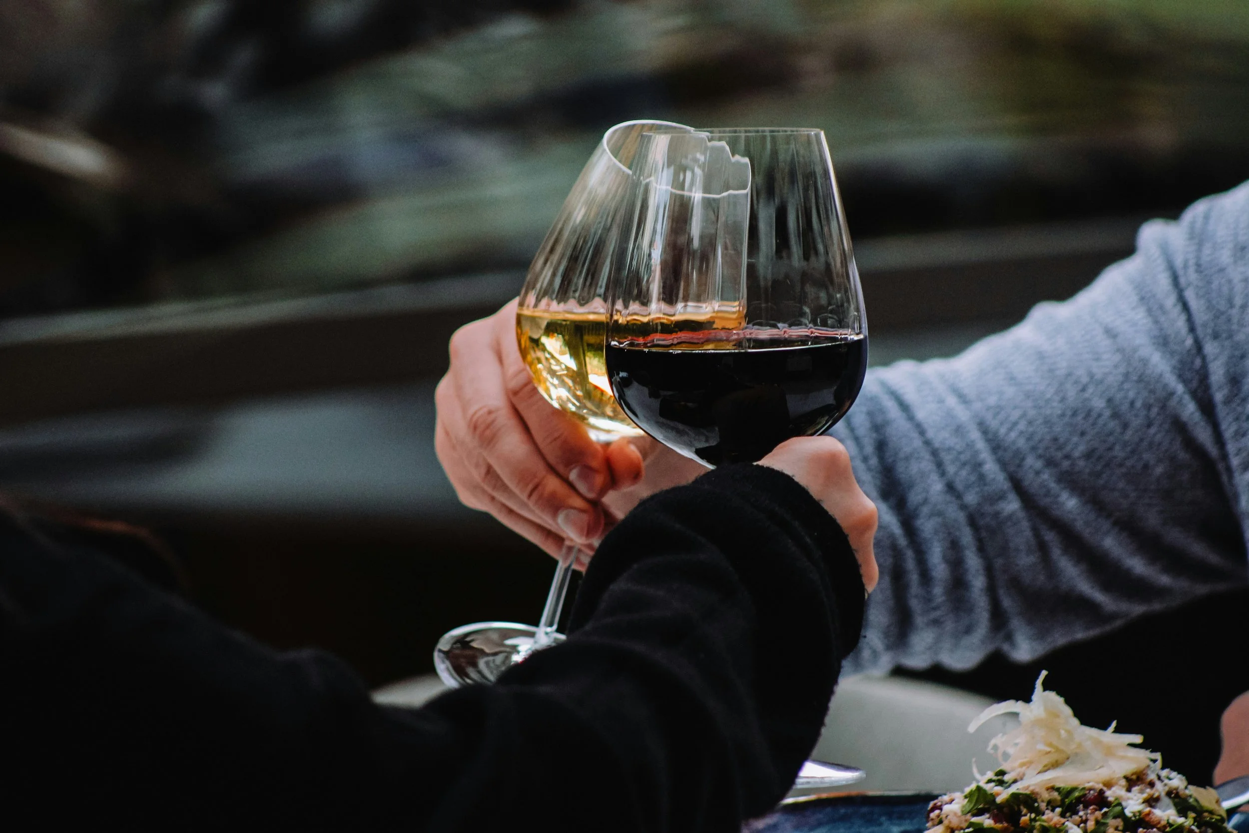 Close-up of a hand holding a glass of white wine next to another hand holding a glass of red wine during a social gathering.
