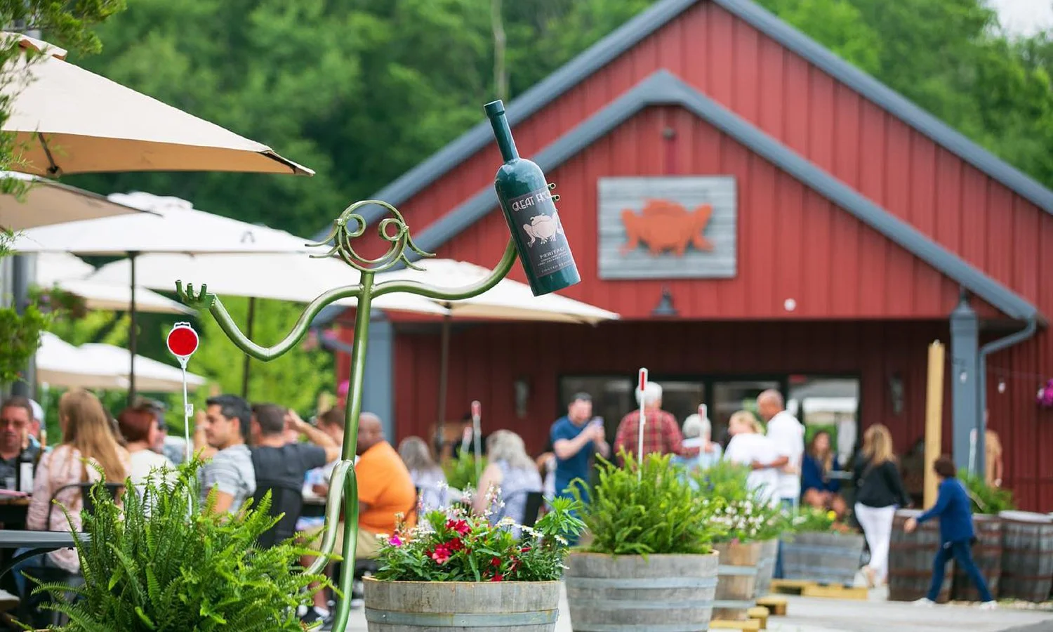 Outdoor restaurant with people dining, potted plants, and a red barn-style building in the background featuring a pig silhouette sign.