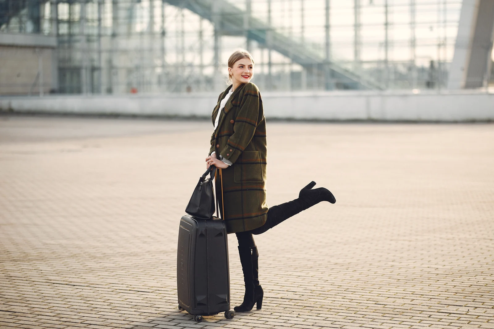 A woman in a long plaid coat, high heels, and black pants standing on an outdoor paved area with luggage and a handbag, smiling and posing with one leg raised behind her.