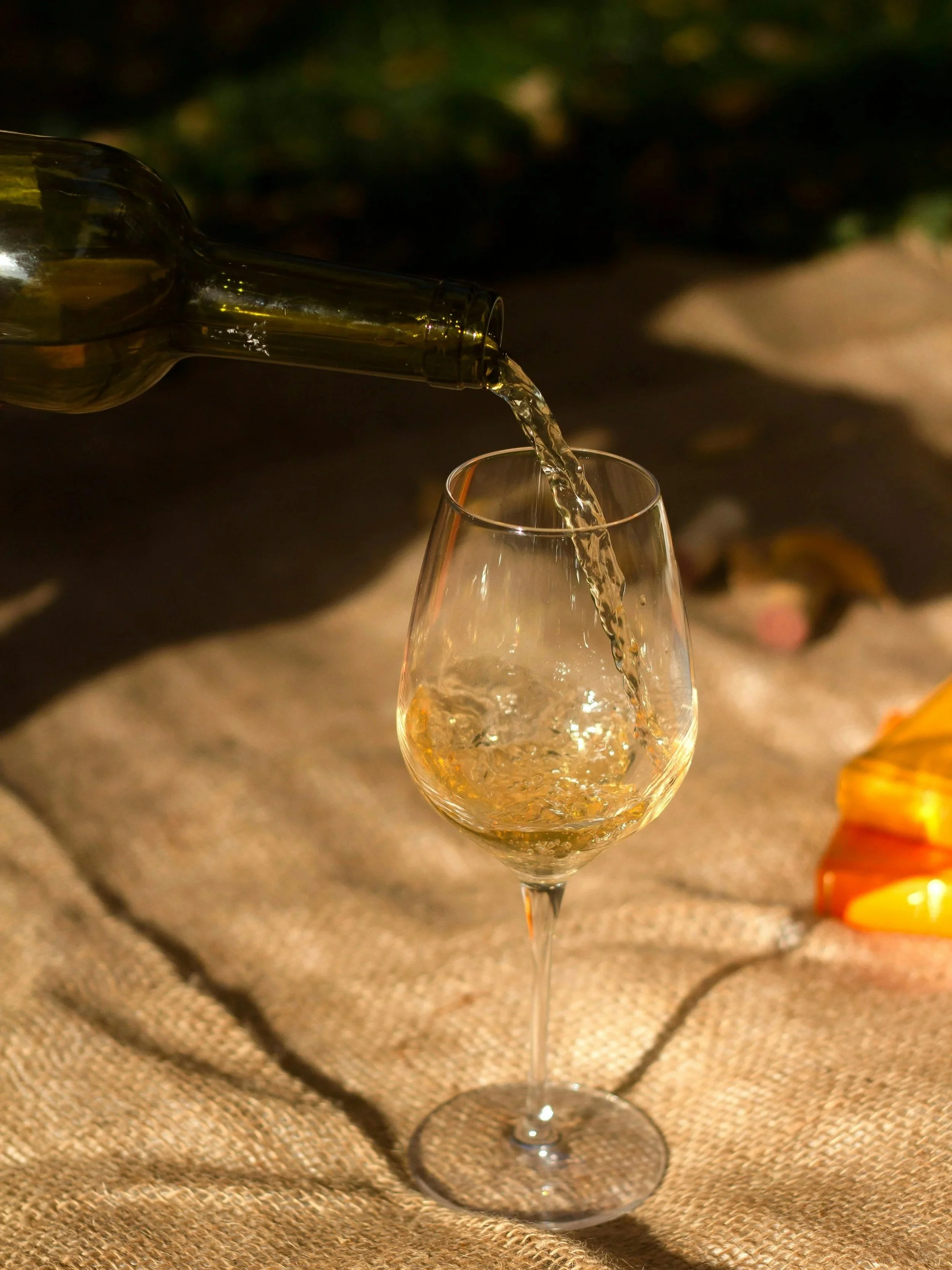 A glass of white wine being filled from a green bottle, set on a rustic fabric surface with autumn leaves in the background.
