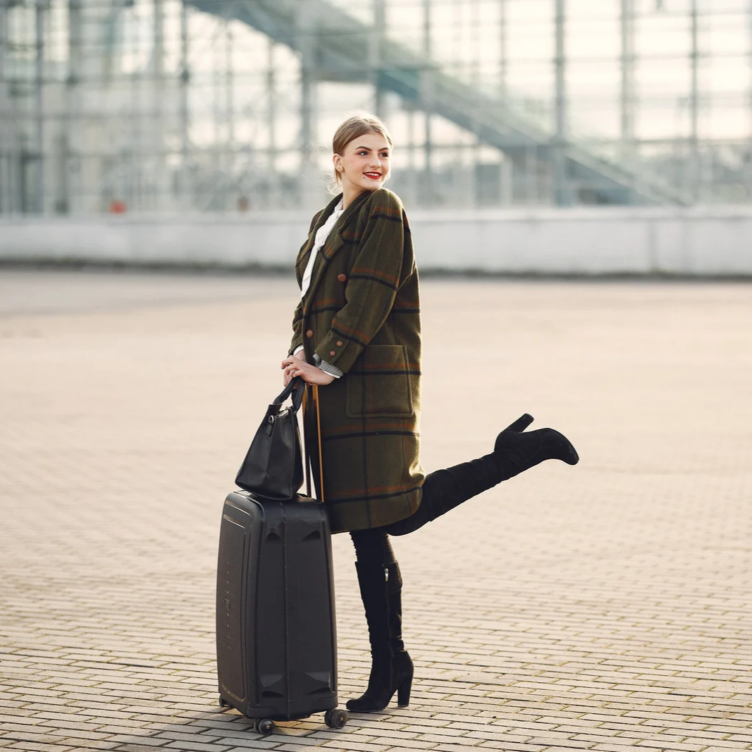 A woman standing at an airport terminal, wearing a green plaid coat, holding a black handbag, with a rolling suitcase, smiling and standing on one leg.