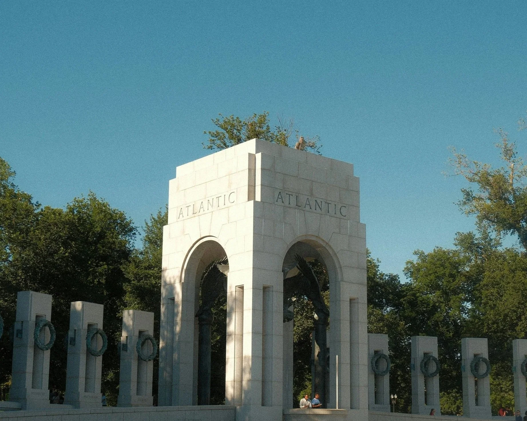 The National World War II Memorial in Washington, D.C., with a large stone archway inscribed with 'Atlantic' and 'Pacific,' featuring bronze sculptures, columns with wreaths, and visitors walking nearby.
