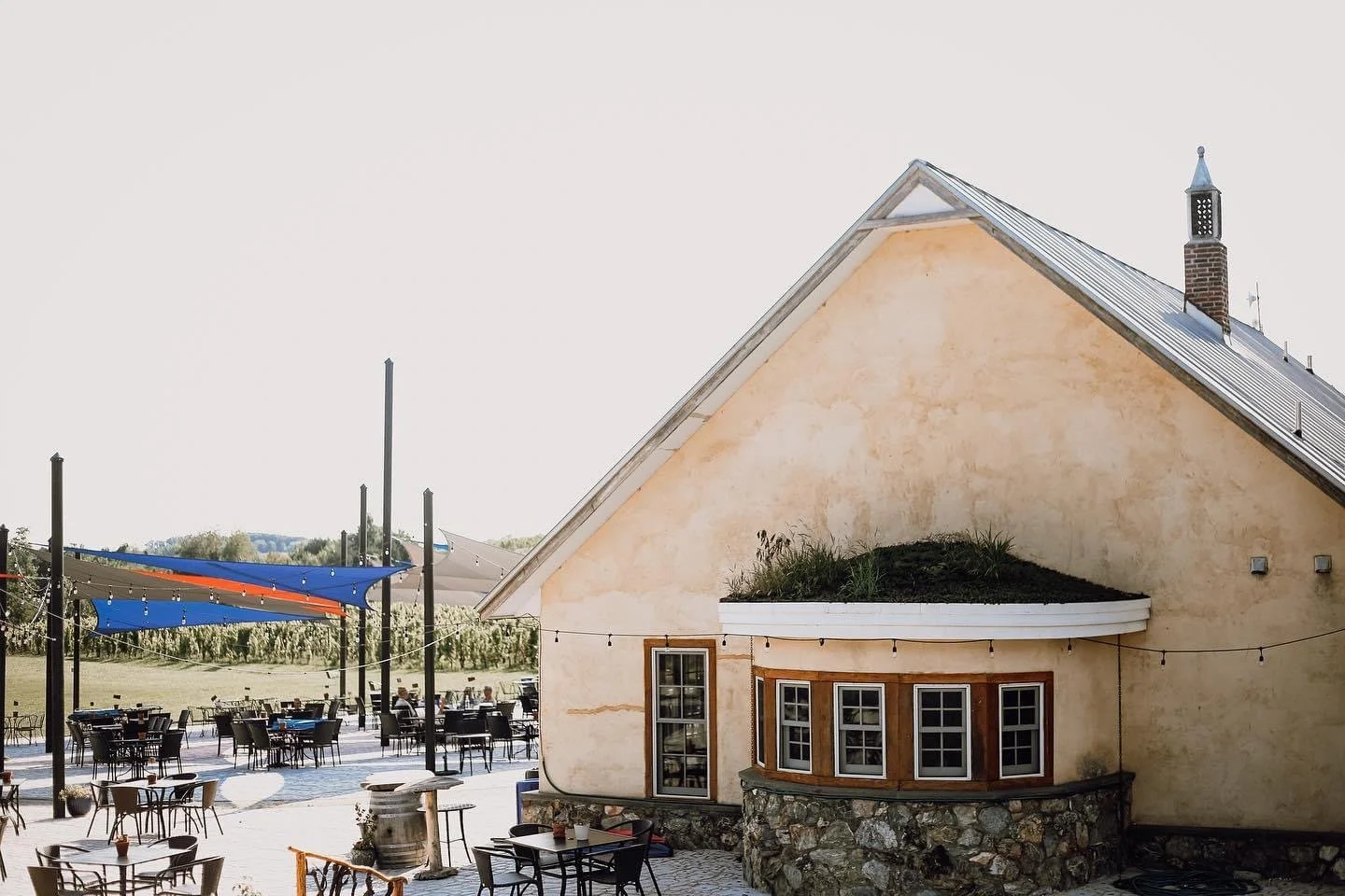 An outdoor patio area with black tables and chairs, some with tables filled with drinks and food, under blue and orange shade sails, next to a small building with a yellow stucco exterior, multiple windows, and a sloped roof with a chimney at the top.