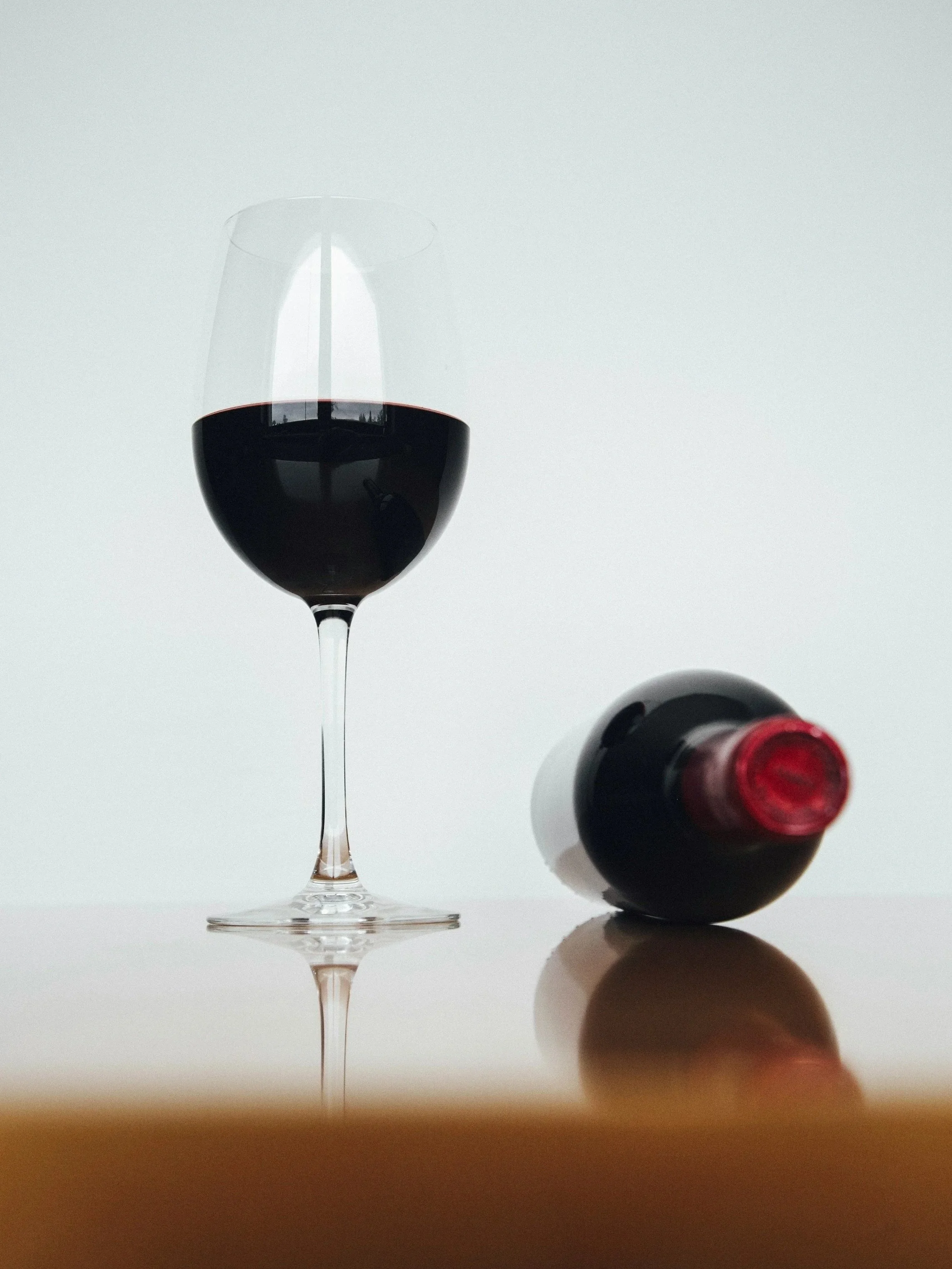 A glass of red wine and a bottle of wine on a reflective surface, with a soft gray background.