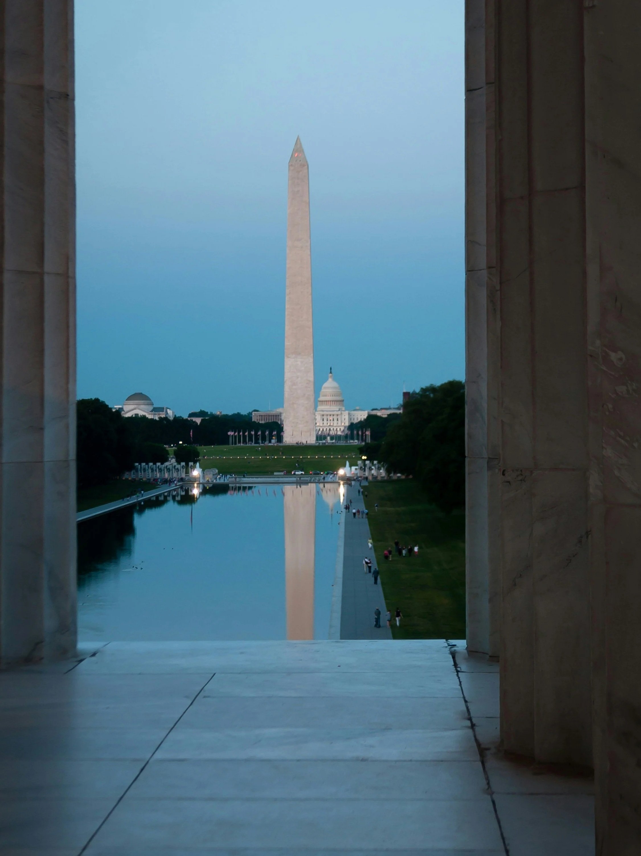 View of the Washington Monument and the United States Capitol from the Lincoln Memorial, framed by marble columns, with a reflecting pool and people walking by in the foreground.