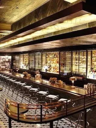 Modern bar counter with bar stools, illuminated shelves with bottles and glassware, black and gold decor, and geometric patterned floor.