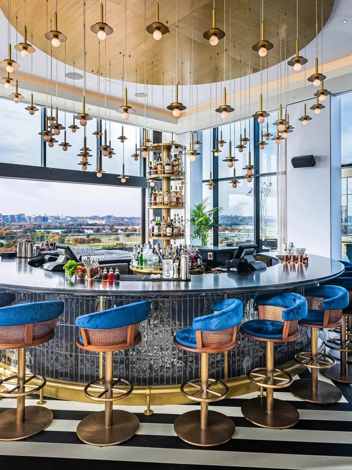 Modern bar with a circular marble countertop, blue velvet chairs, and a central shelving unit with liquor bottles. Large windows reveal a cityscape and open sky. Pendant lights hang from a gold ceiling, and a potted plant adds greenery to the space.