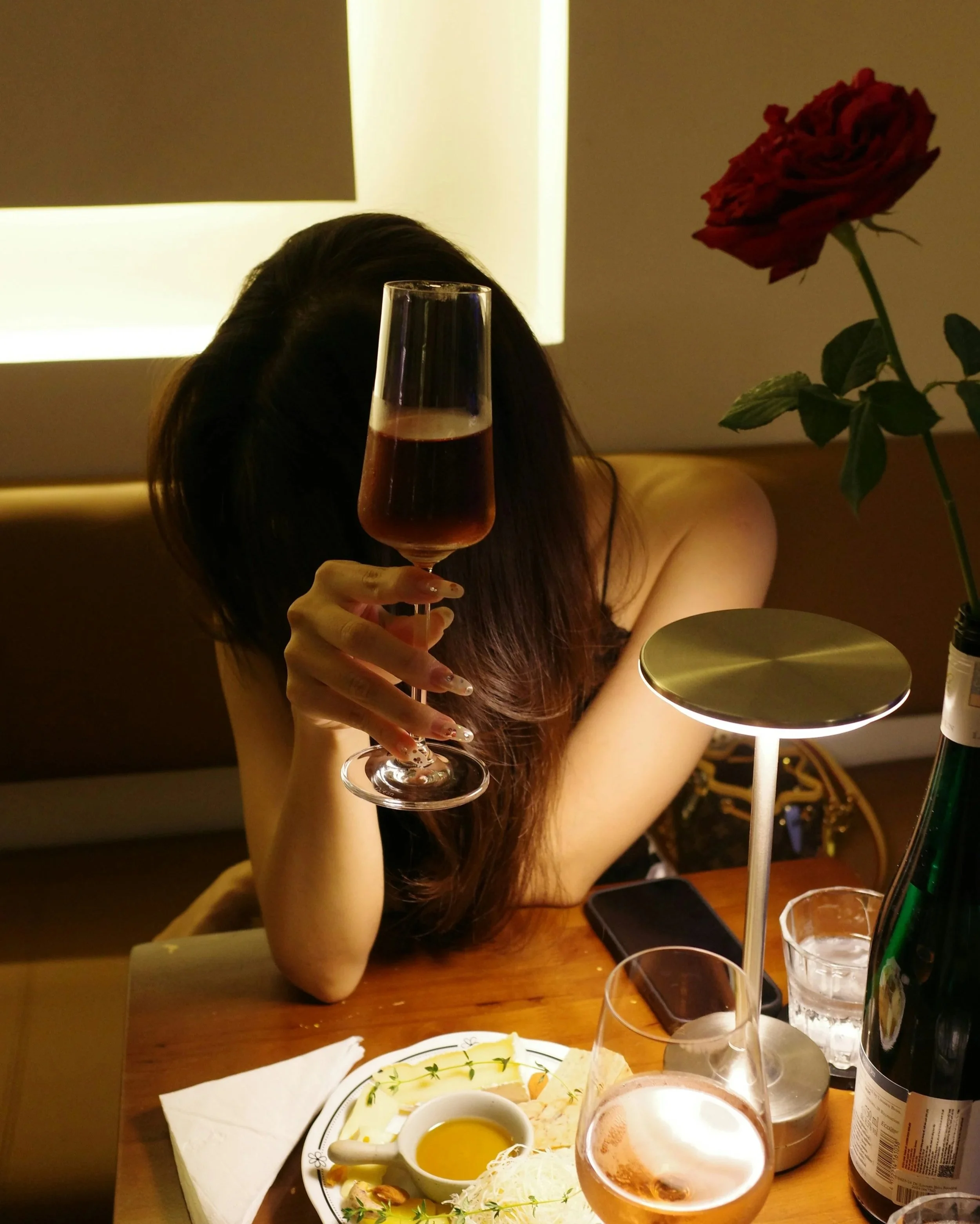 A woman with long dark hair, holding a glass of rosé wine, sitting at a dining table with plates of food, glasses, and a bottle of rosé wine in the background, with a red rose on the table.