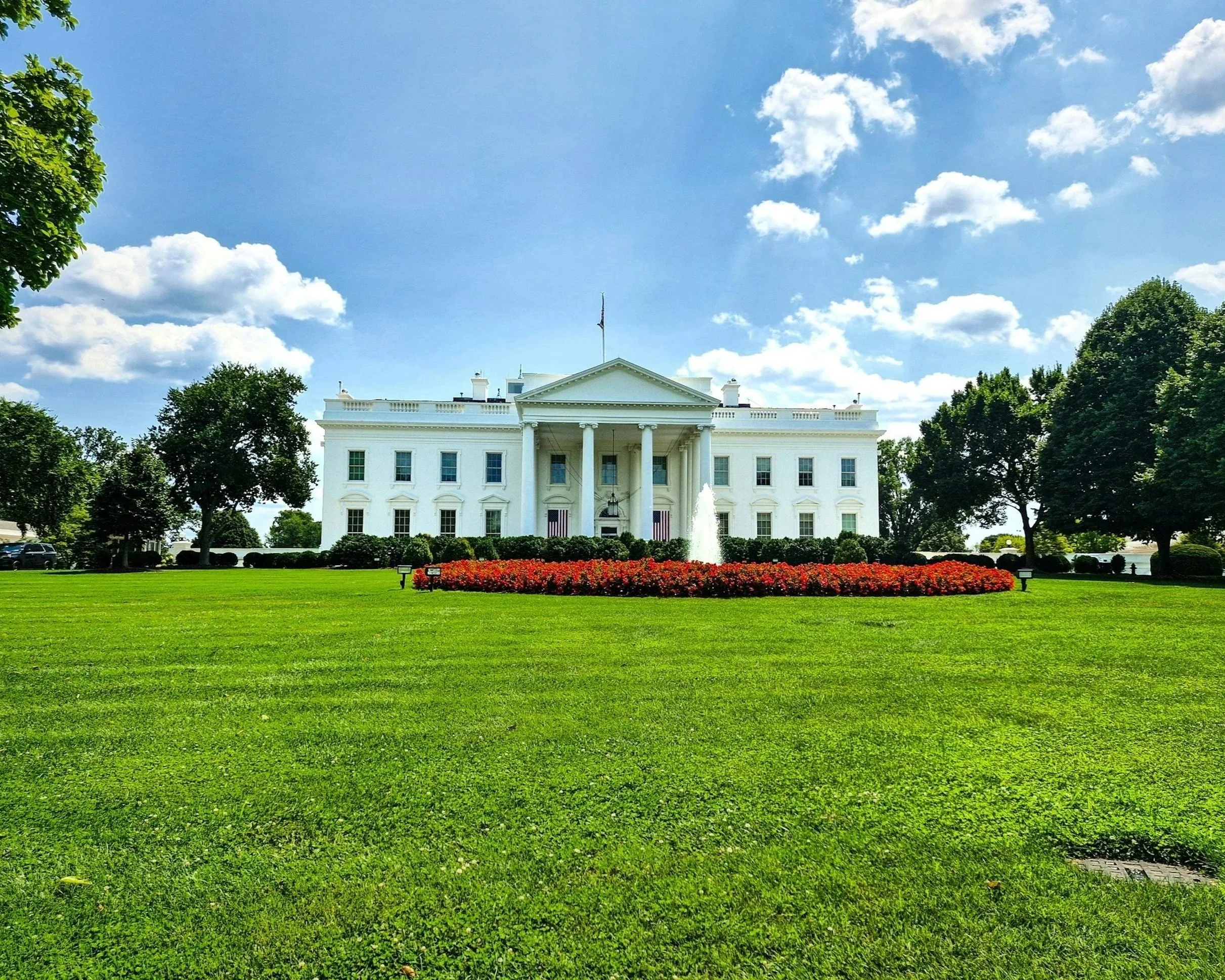 The White House, a neoclassical mansion with a central portico, columns, and an American flag on top, surrounded by green trees and a lawn under a partly cloudy sky.