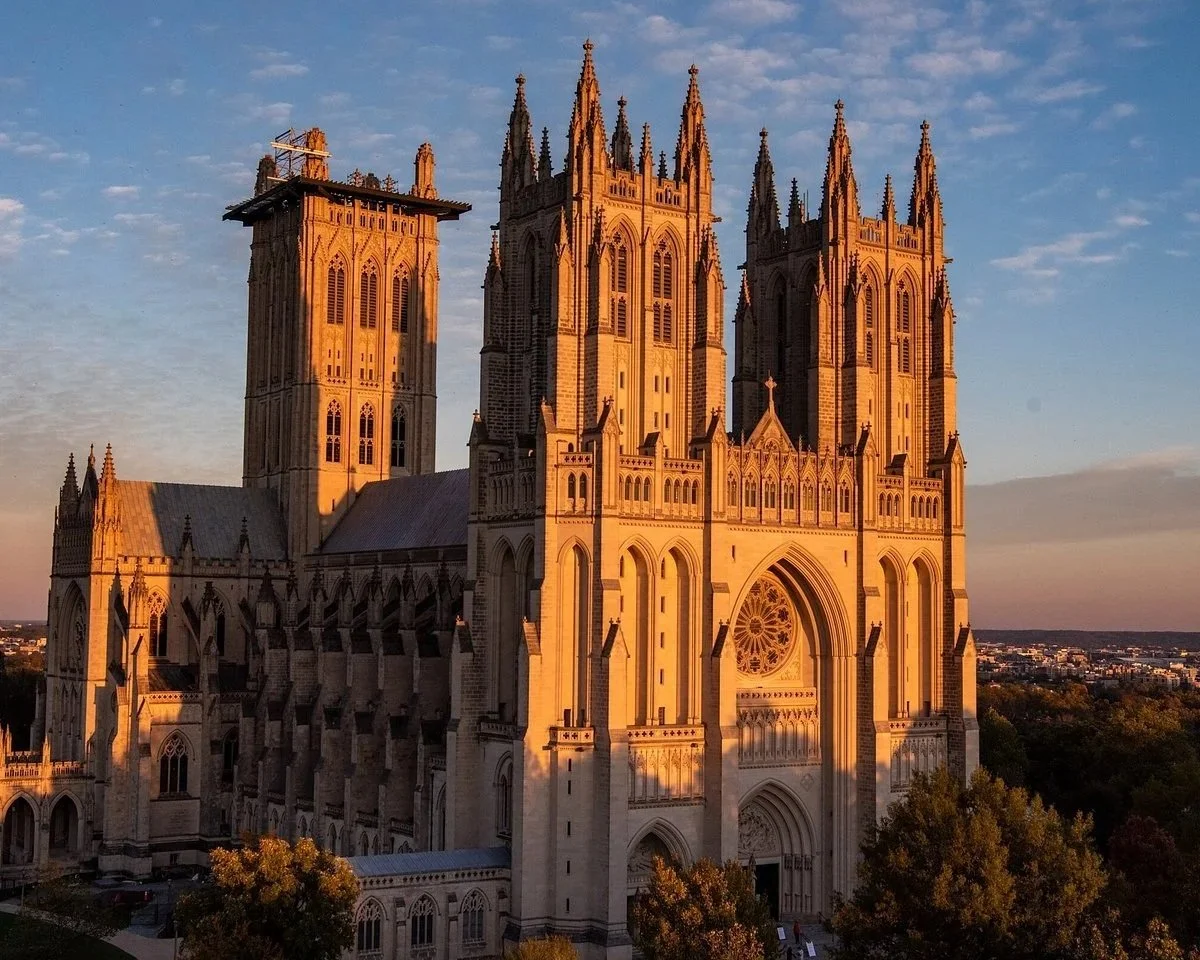 A large Gothic cathedral with multiple towers and intricate architectural details, illuminated by the warm light of the setting sun, with trees in the foreground and a cityscape in the distance.