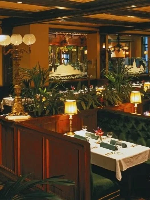 Elegant restaurant interior with dark wood furnishings, pendant lamps, and lush green plants, set with white tablecloths, wine glasses, and floral centerpieces.