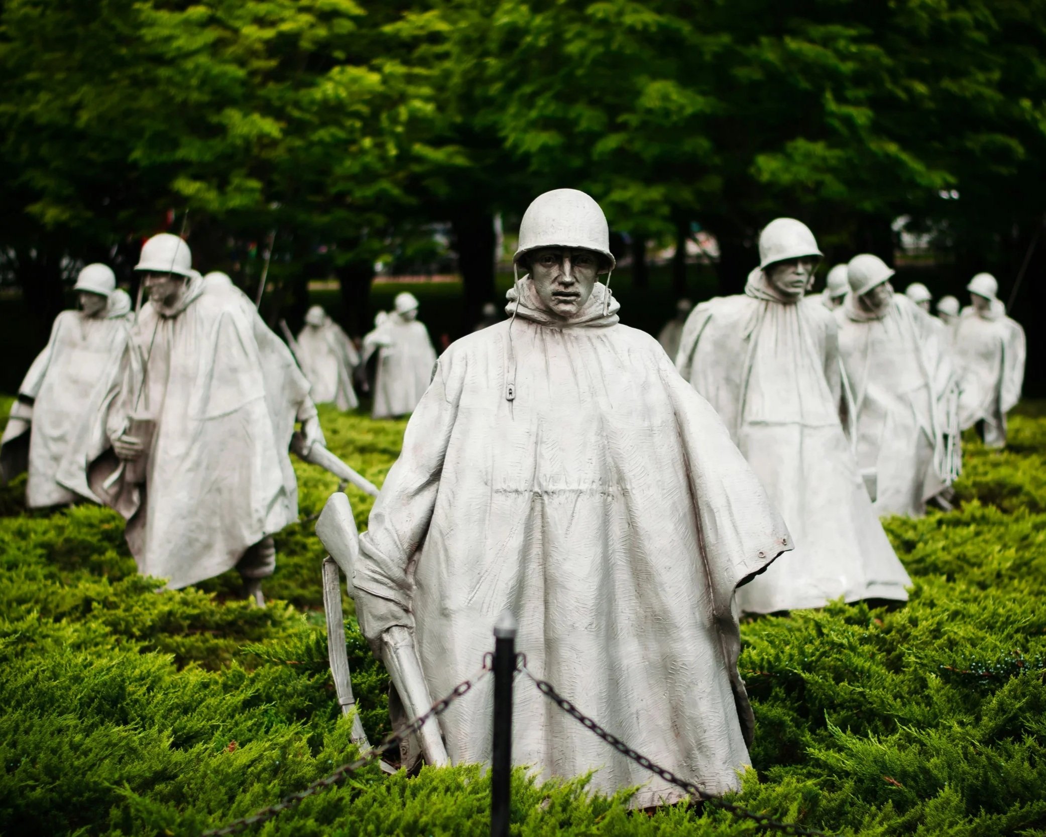 A memorial with bronze statues of soldiers wearing helmets and cloaks, standing in a park with green trees in the background, surrounded by a chain fence.
