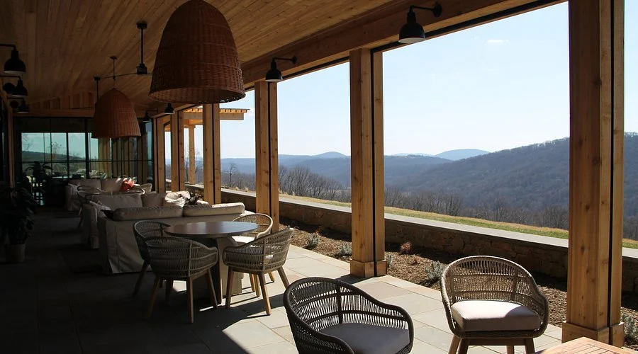 Indoor patio area with wooden ceiling, hanging wicker lamps, and patio furniture including wicker chairs and a sofa, overlooking a scenic mountain view through large open windows.