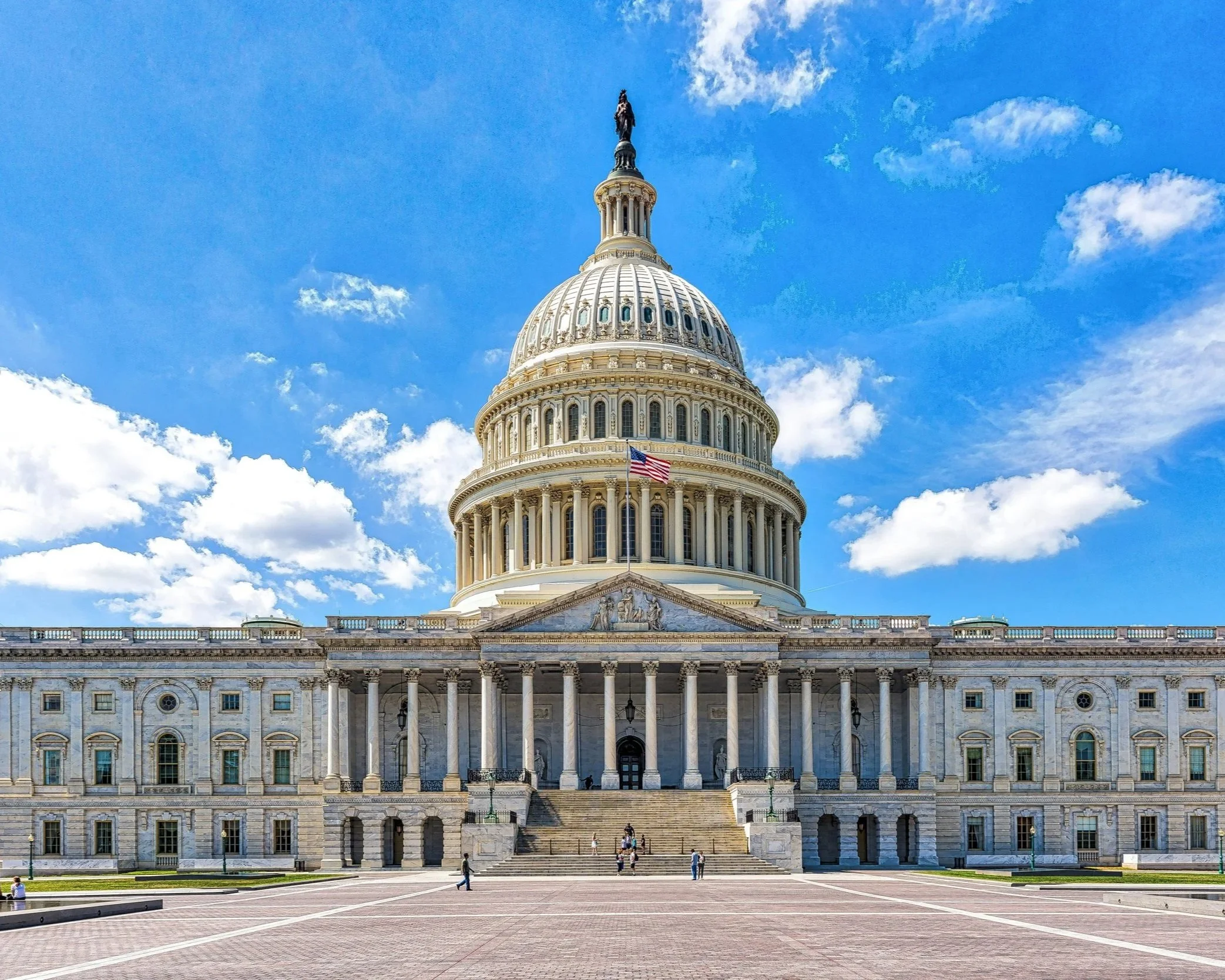 The United States Capitol building with a clear blue sky and scattered clouds in the background. The front steps and façade of the monument are visible with a few people walking in the foreground, and an American flag displayed prominently.