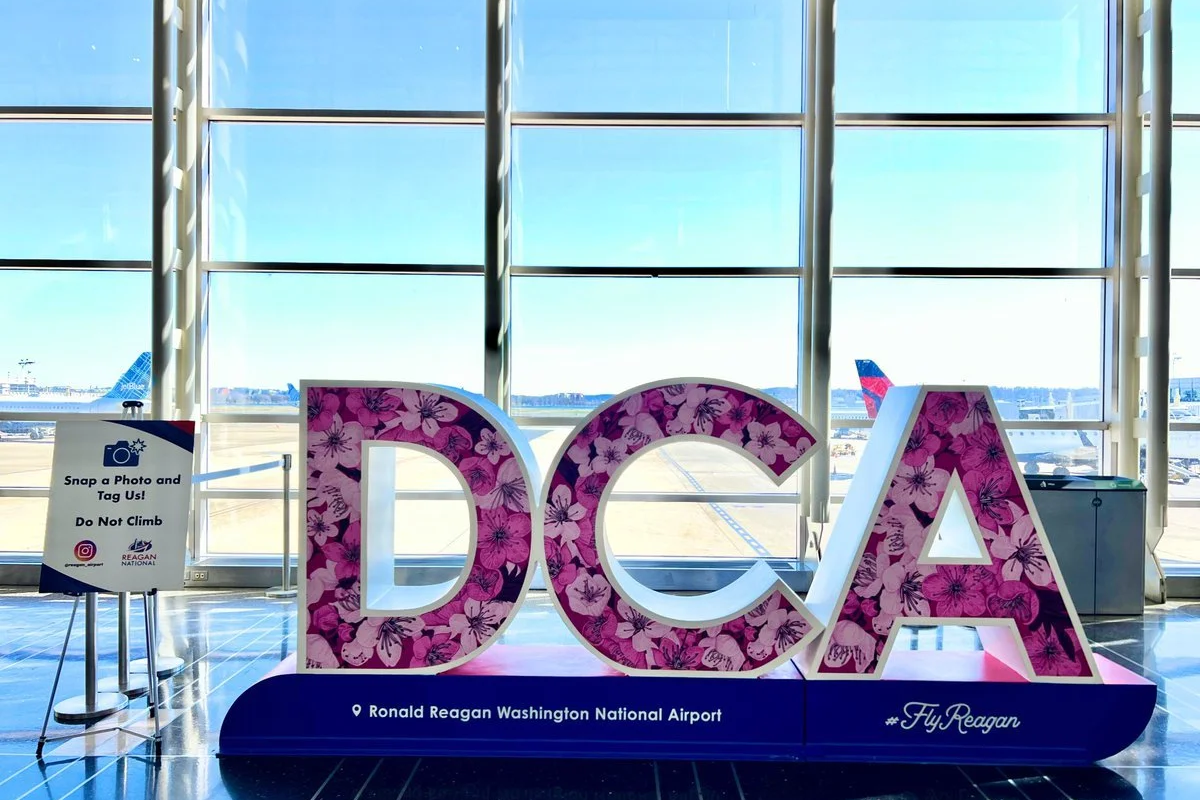 Decorative sign at Ronald Reagan Washington National Airport spelling out "DCA" with pink floral patterns, with airplanes visible through large windows in the background.