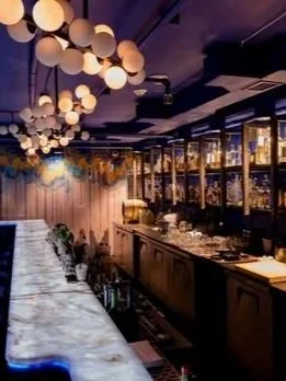Elegant bar with a marble countertop, illuminated globe pendant lights, and large windows showing a cityscape at night.