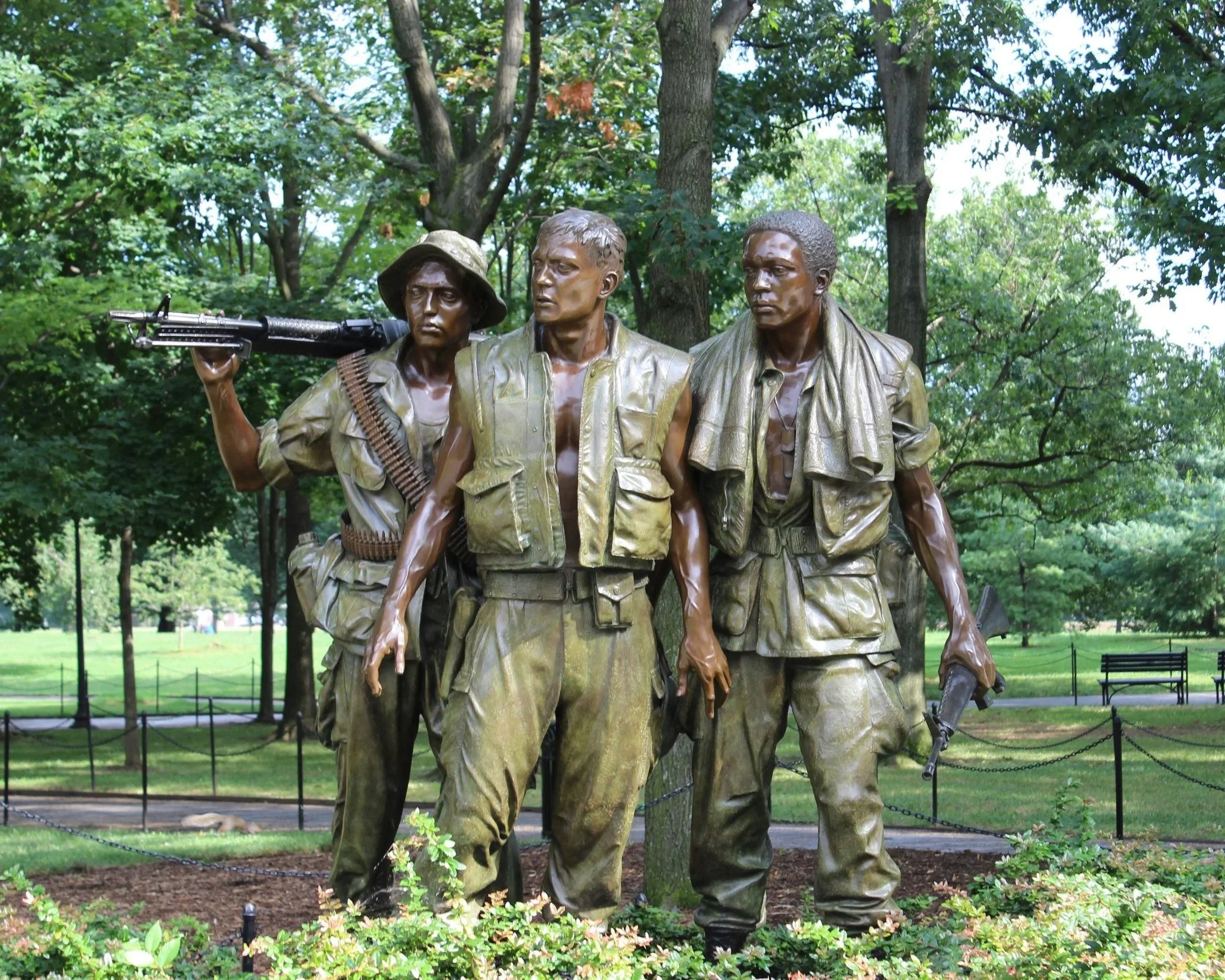 Bronze statues of four soldiers standing outdoors in a park, one holding a rifle, surrounded by trees and greenery.