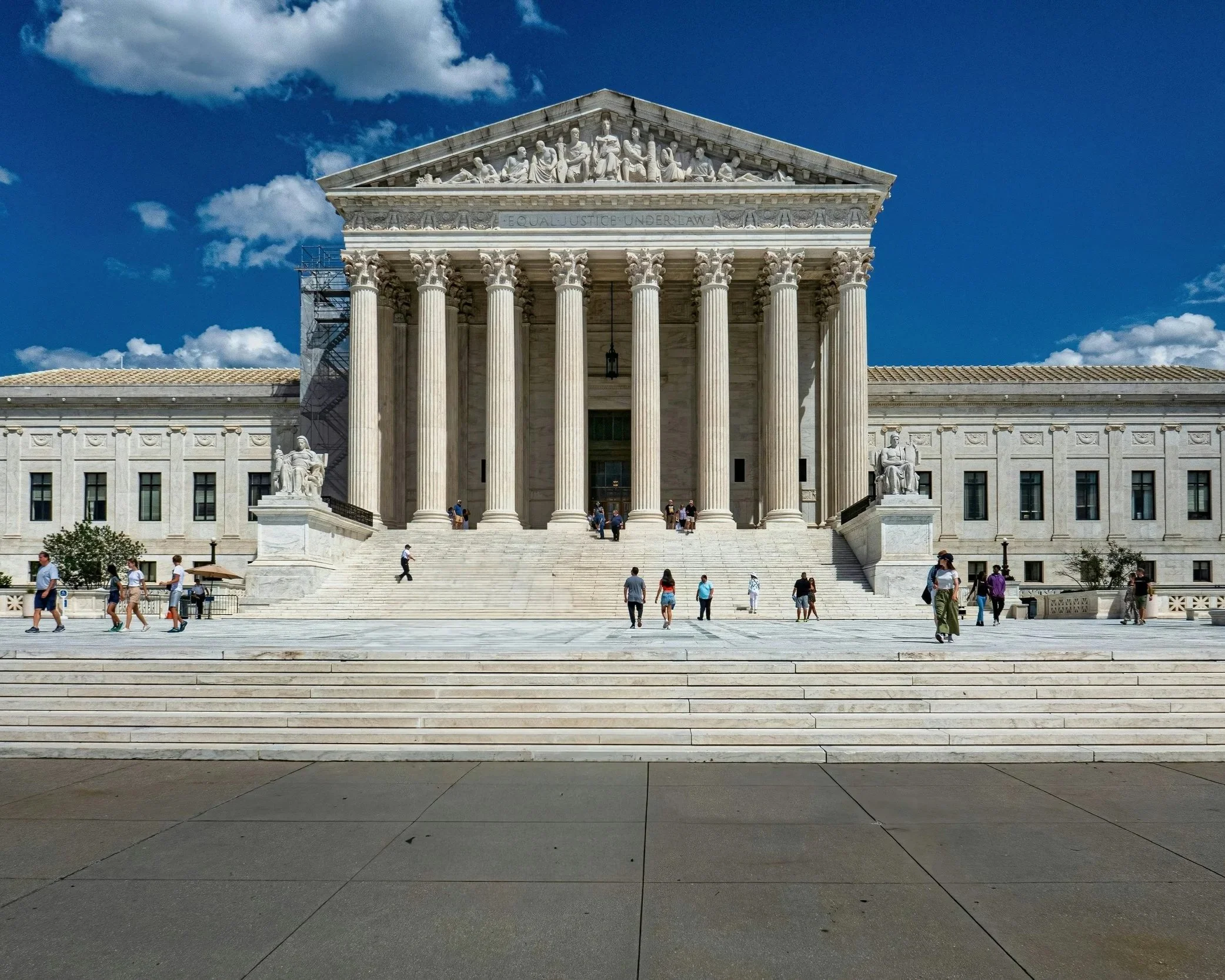 The United States Supreme Court building, featuring classical architecture with large columns and sculptures, seen from the steps with visitors walking.
