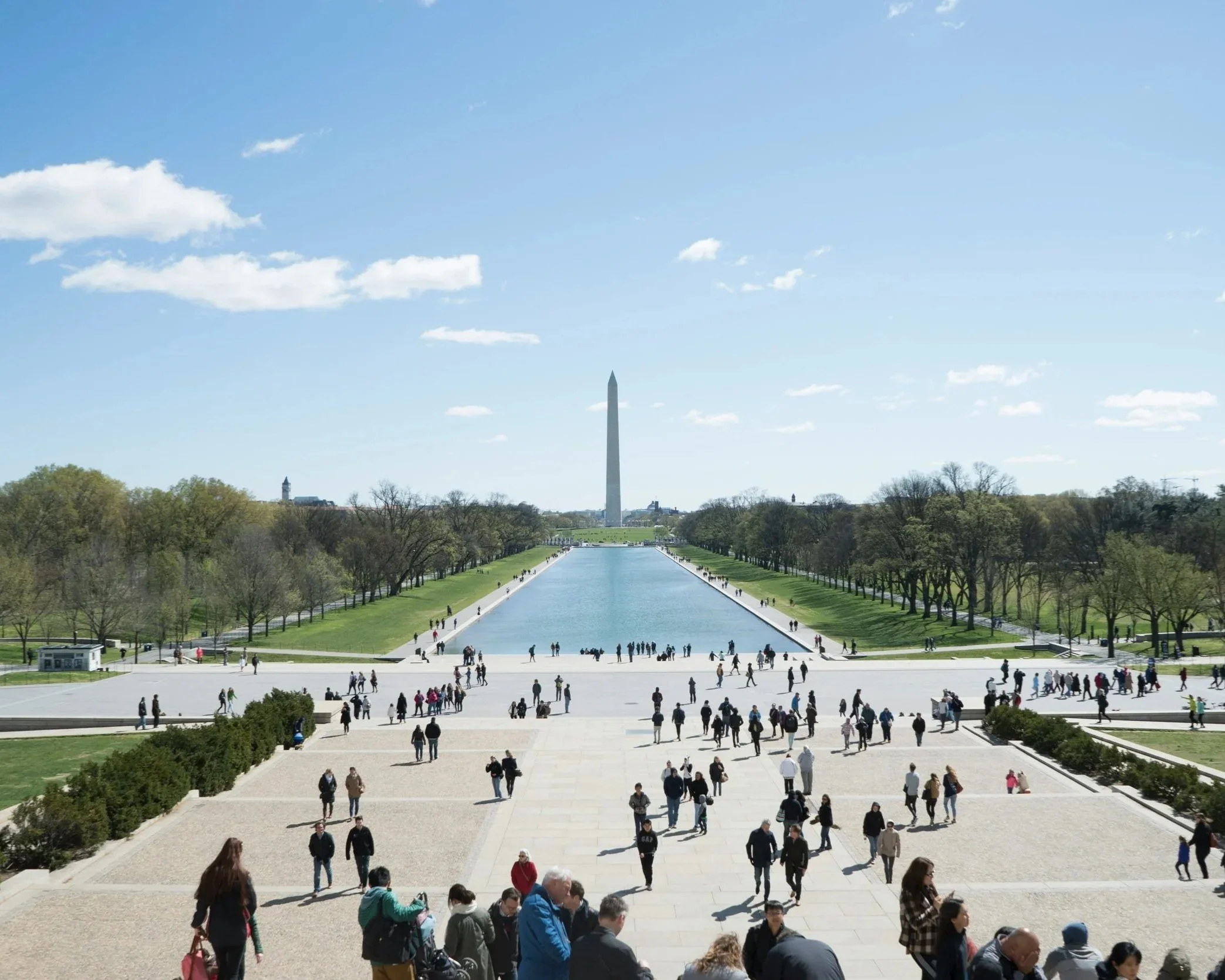View of the National Mall in Washington, D.C., featuring the Washington Monument in the background, a reflecting pool, and people walking and gathering on the steps and grass.