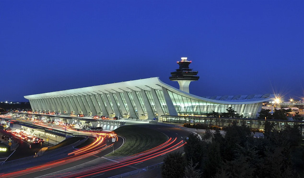 Nighttime view of a modern airport terminal with a distinctive curved roof and a control tower in the background, with streaks of car lights on the road in front.