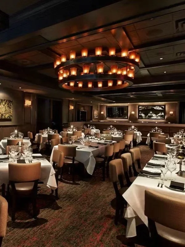 Elegant restaurant dining room with round tables set with white tablecloths, glassware, and folded napkins, illuminated by a large circular chandelier with lit candles, dark wood paneling, and a bar area in the background.